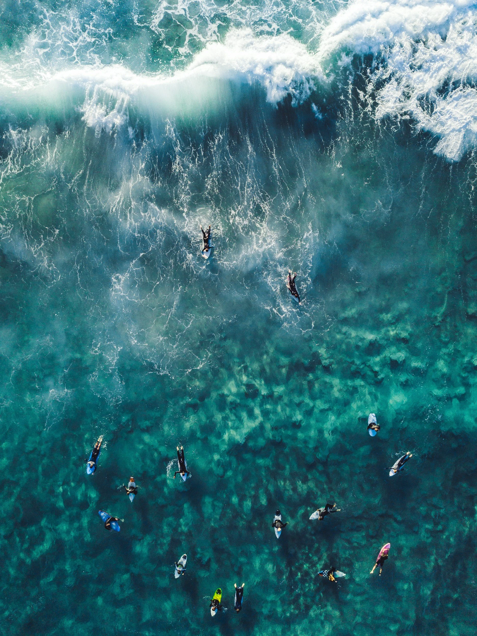 An aerial view of multiple surfers riding a large ocean wave in clear blue water.