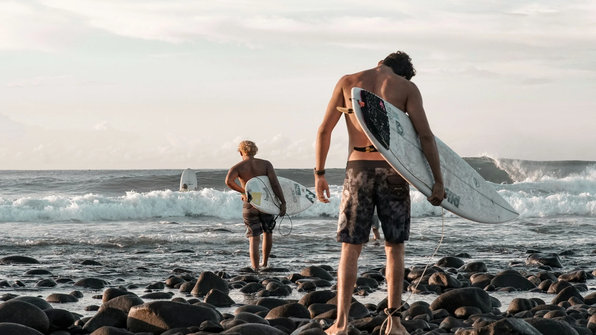 Two young men with surfboards standing on a rocky shoreline, facing the ocean with small waves, during a cloudy day.