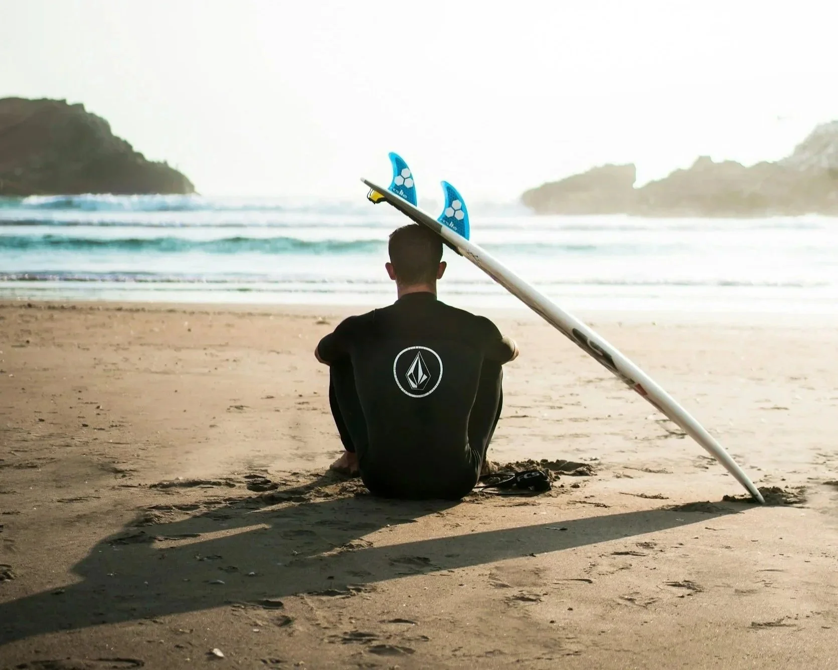 A person sitting on the beach facing the ocean with a surfboard resting on their head.