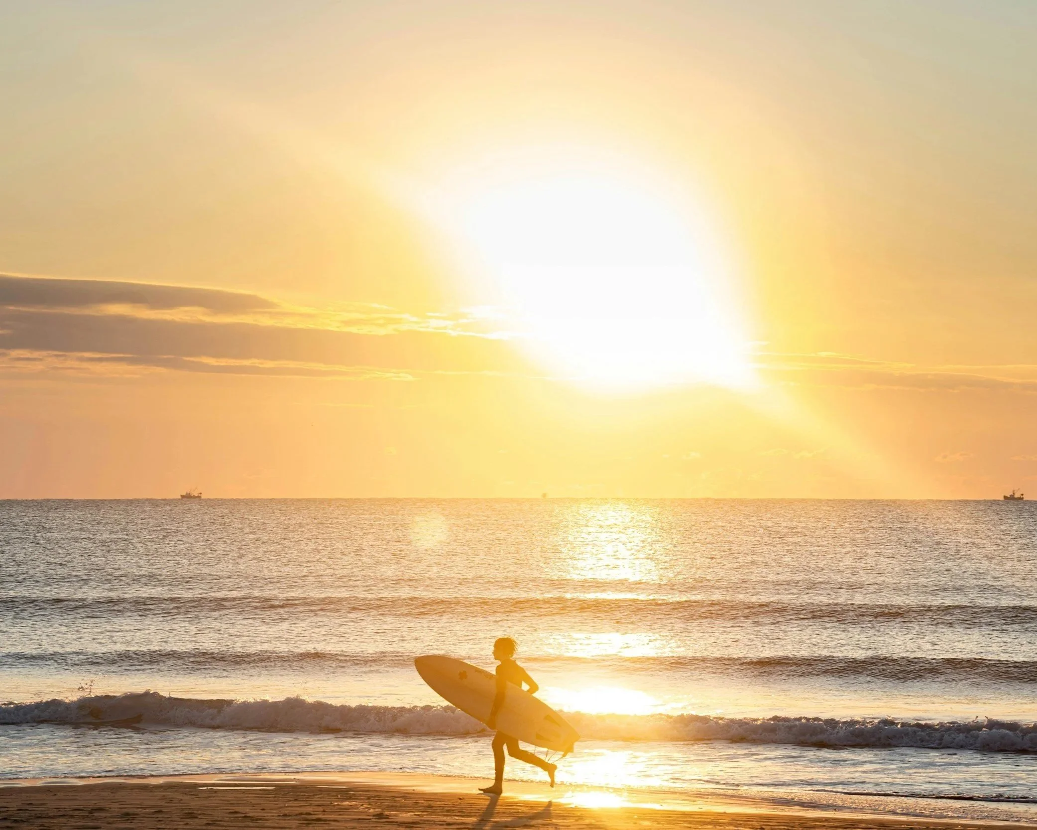 A person stands on the beach holding a surfboard at sunset, with the sun low in the sky, casting a golden glow over the ocean and cliffs in the distance.