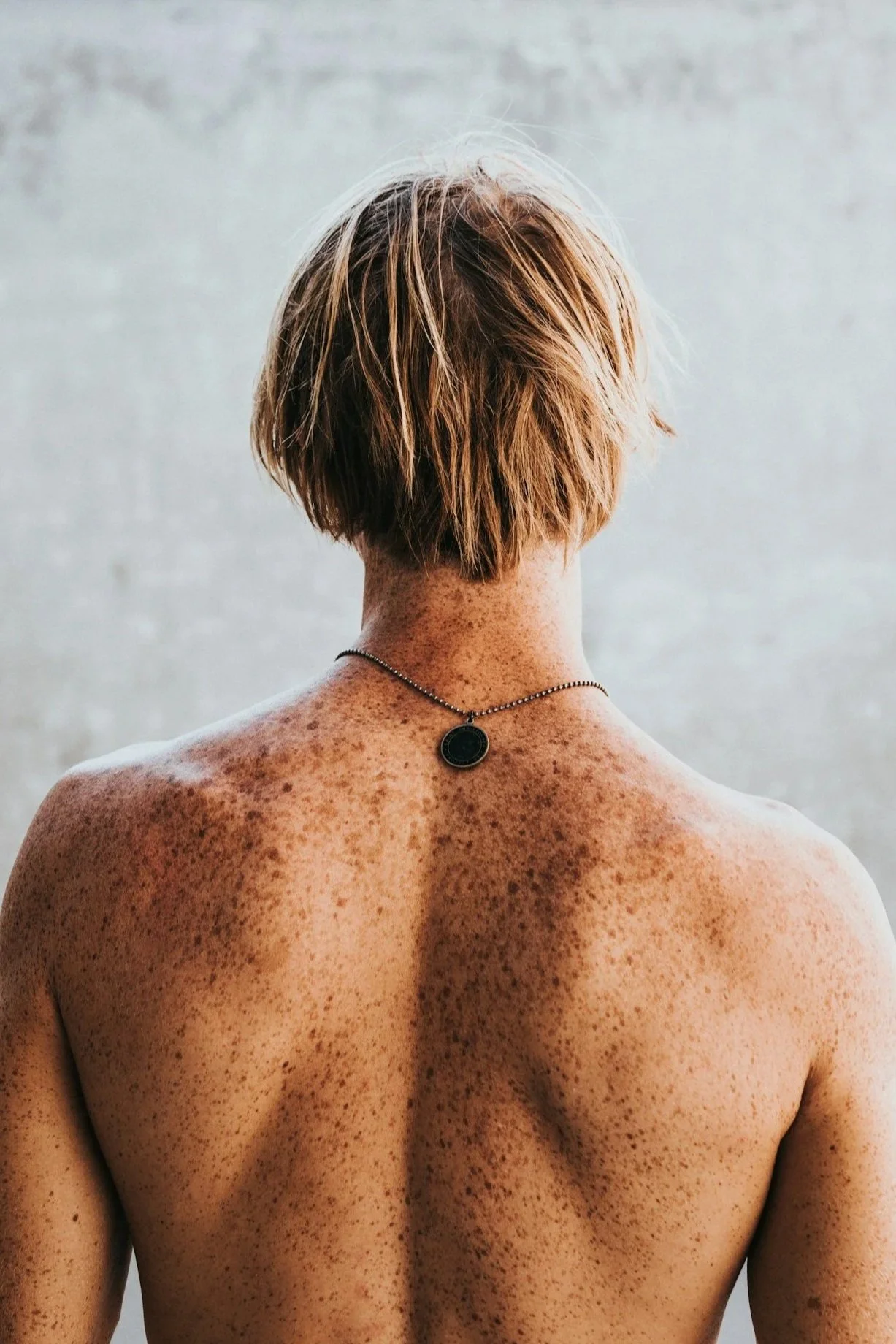 Back view of a person with short, light brown hair and freckled skin, wearing a black pendant necklace.