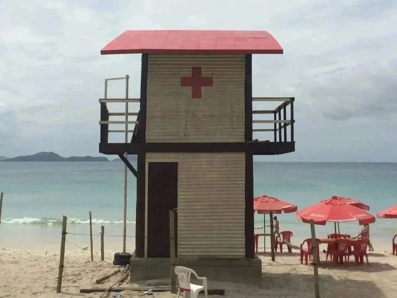 A lifeguard tower with a red cross on its front, located on a beach with red umbrellas and chairs, and a person walking near the water with an island in the background.