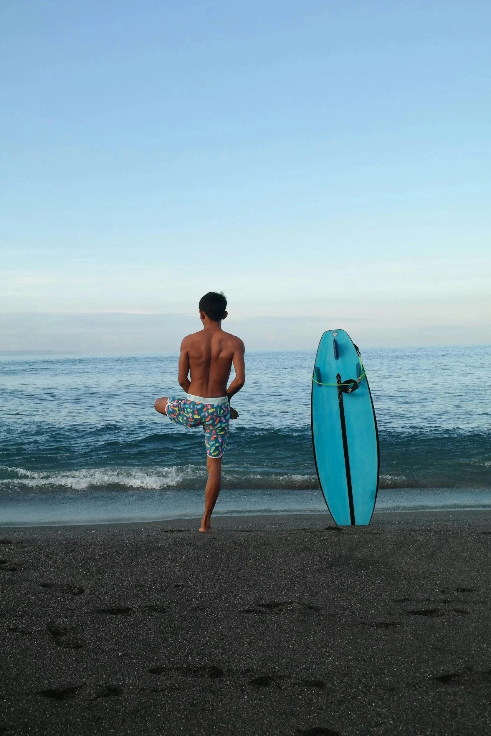A young boy standing on a black sand beach, facing the ocean, wearing colorful swim trunks, with one leg raised in a yoga pose. A blue surfboard stands upright next to him. The sky is clear, with calm waves in the background.