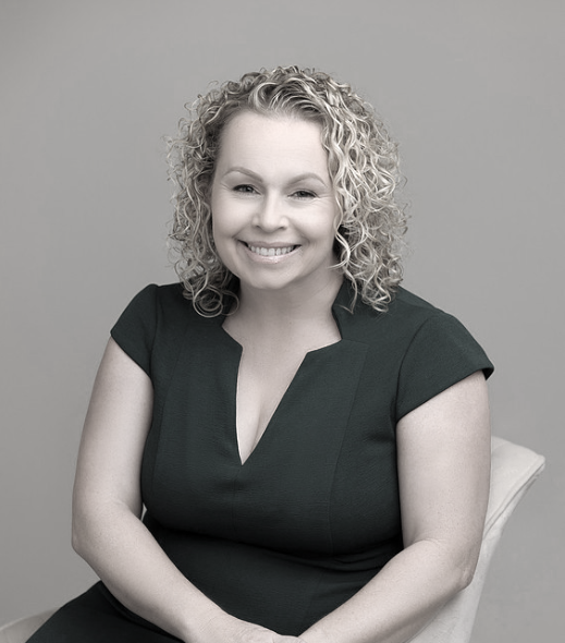 A woman with curly blonde hair smiling while sitting on a chair against a plain background. Founder second mother to many and lover of all things good for children