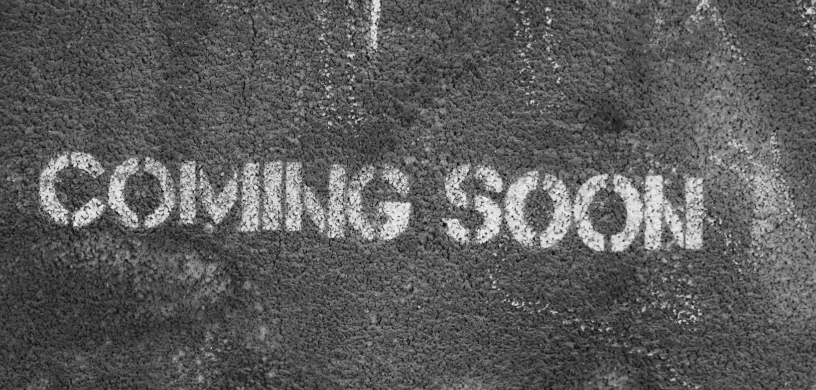Word 'COMING SOON' written with white powder on a dark granular surface.