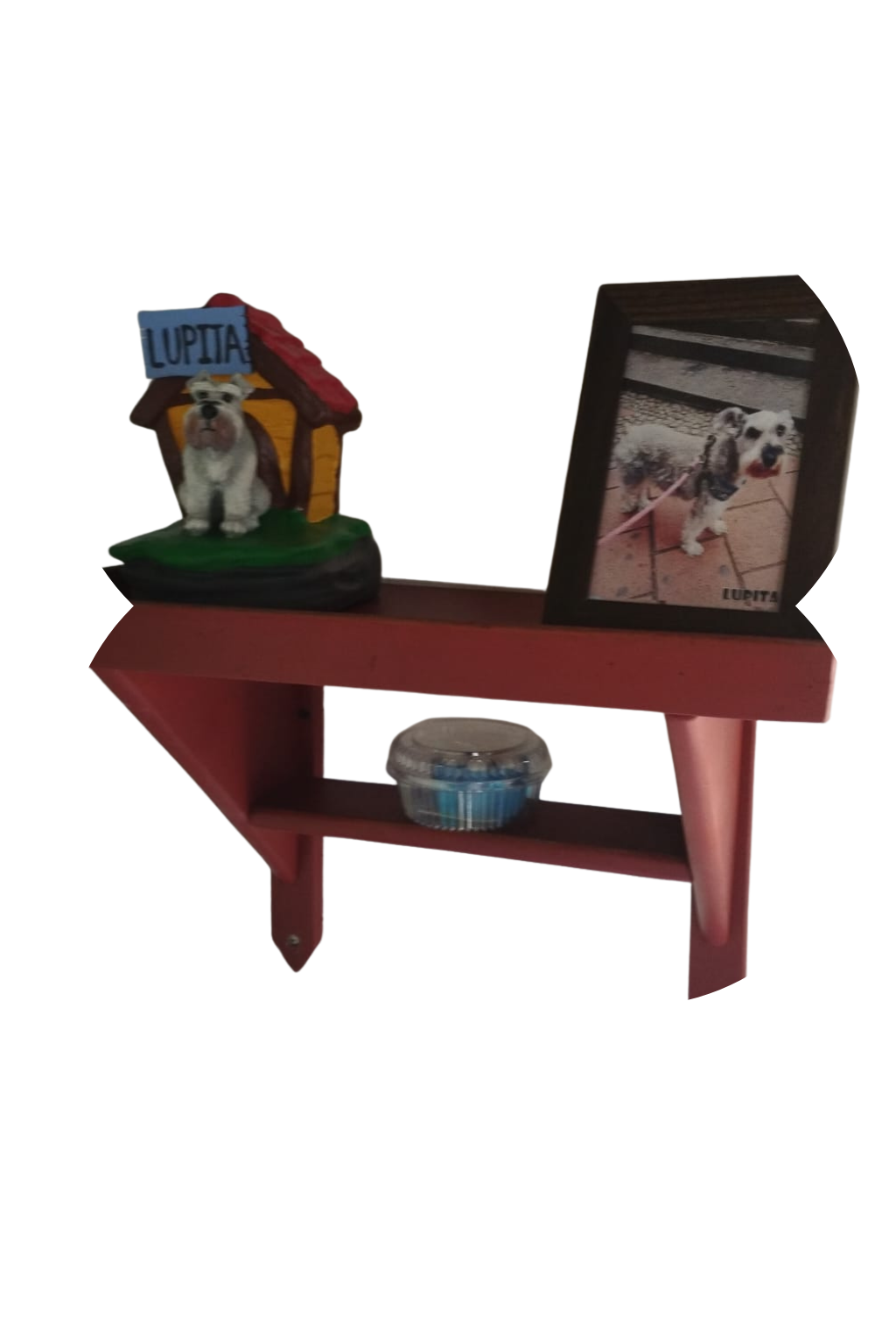 Foto de una mesa de color rojo con una figura decorativa de un perro en una casita y un marco con una foto de un perro llamado Lupita, en un fondo blanco.