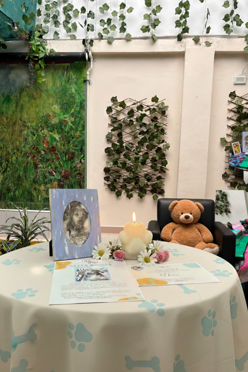 Mesa decorada con foto y descripción de un perro, vela encendida rodeada de flores, en un ambiente con paredes adornadas con plantas, una silla con un oso de peluche