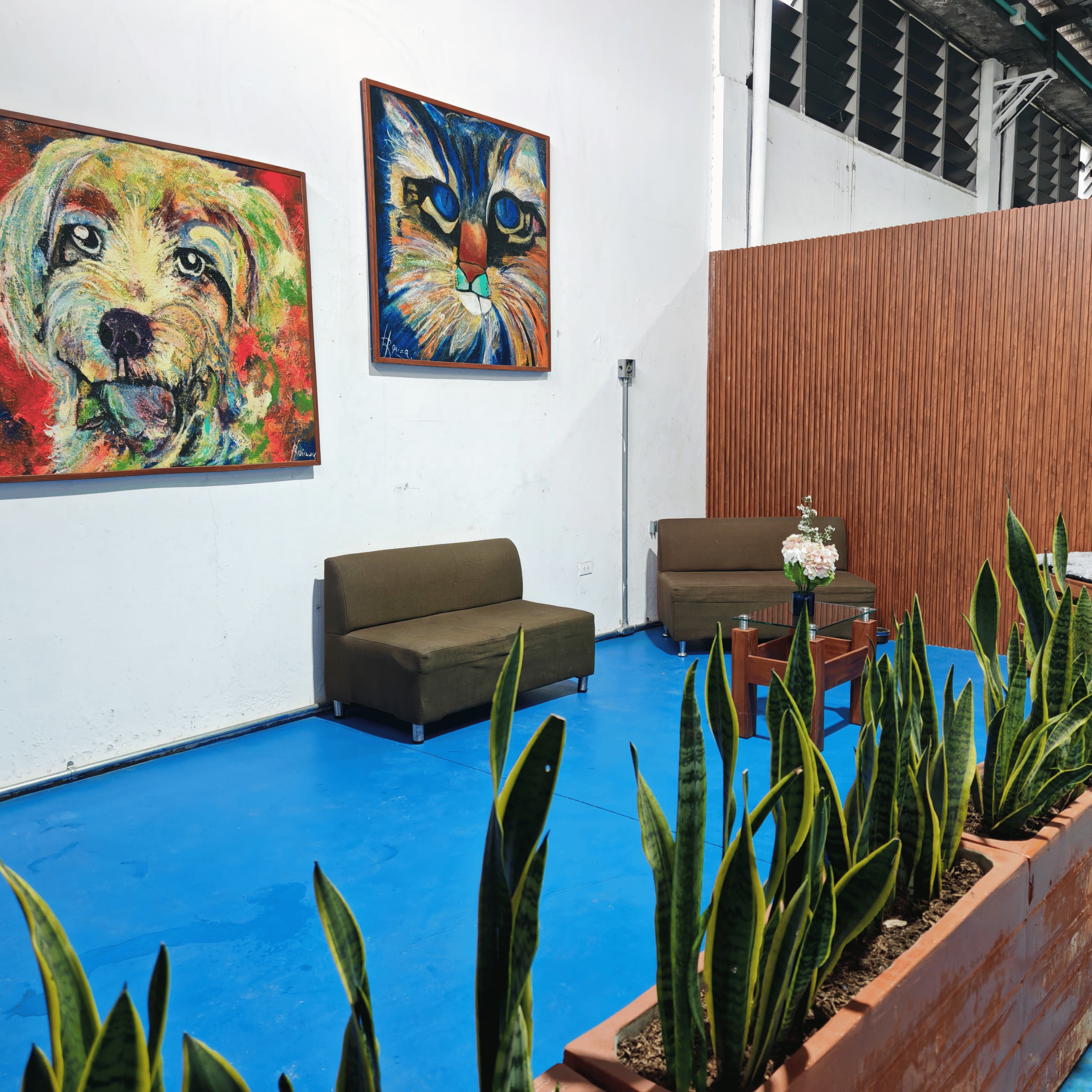 Sala de exposición con dos sofás verdes, una mesa de café con flores, plantas frente a la pared blanca con dos pinturas de animales, una de perro y otra de gato, fondo con pared de madera y estructura de metal en el techo.