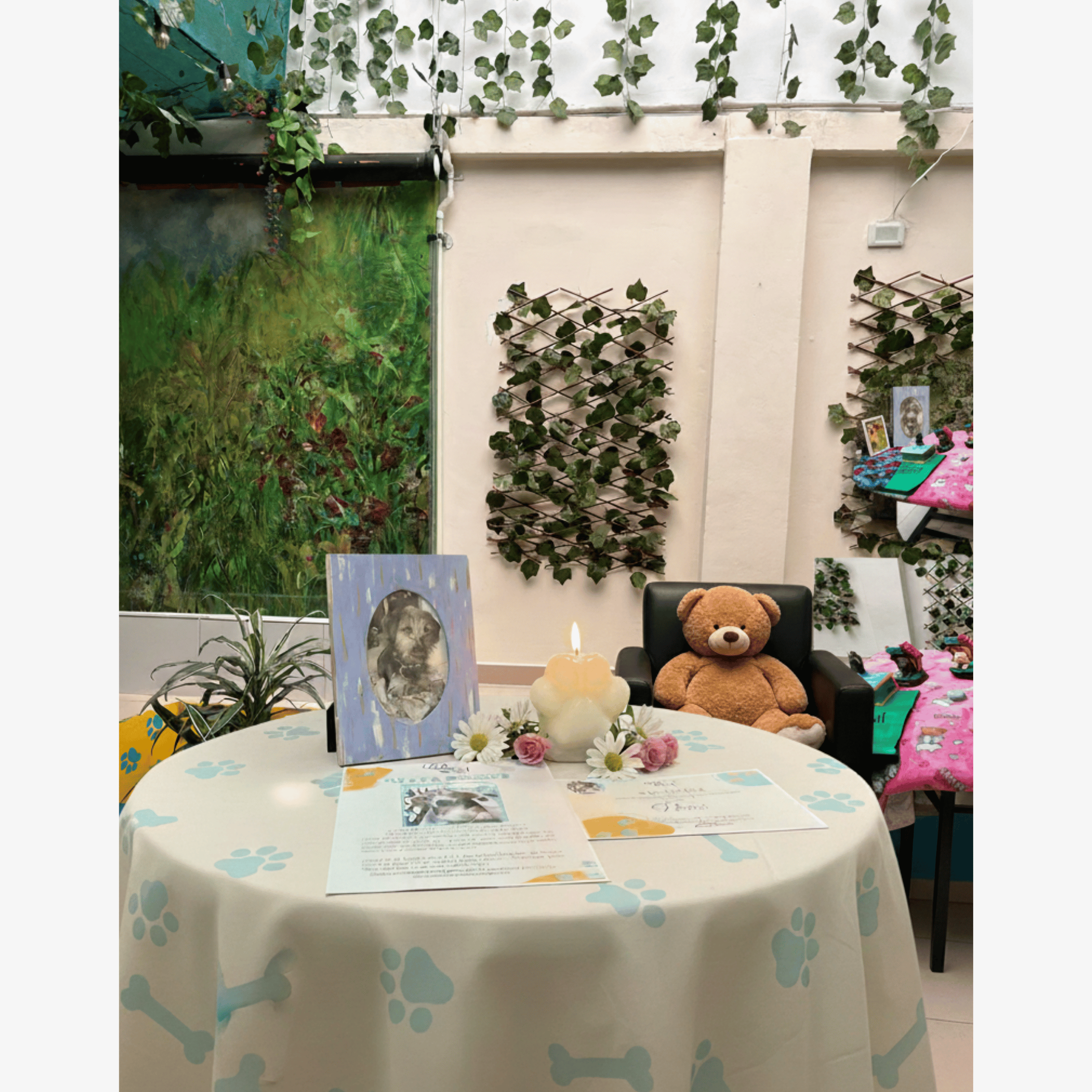 Mesa con fotos de un perro, flores, un libro y un vela con forma de corazón con llama, rodeada de un ambiente decorativo con plantas y paredes con enrejados de hojas verdes, y en el fondo un peluche de oso.