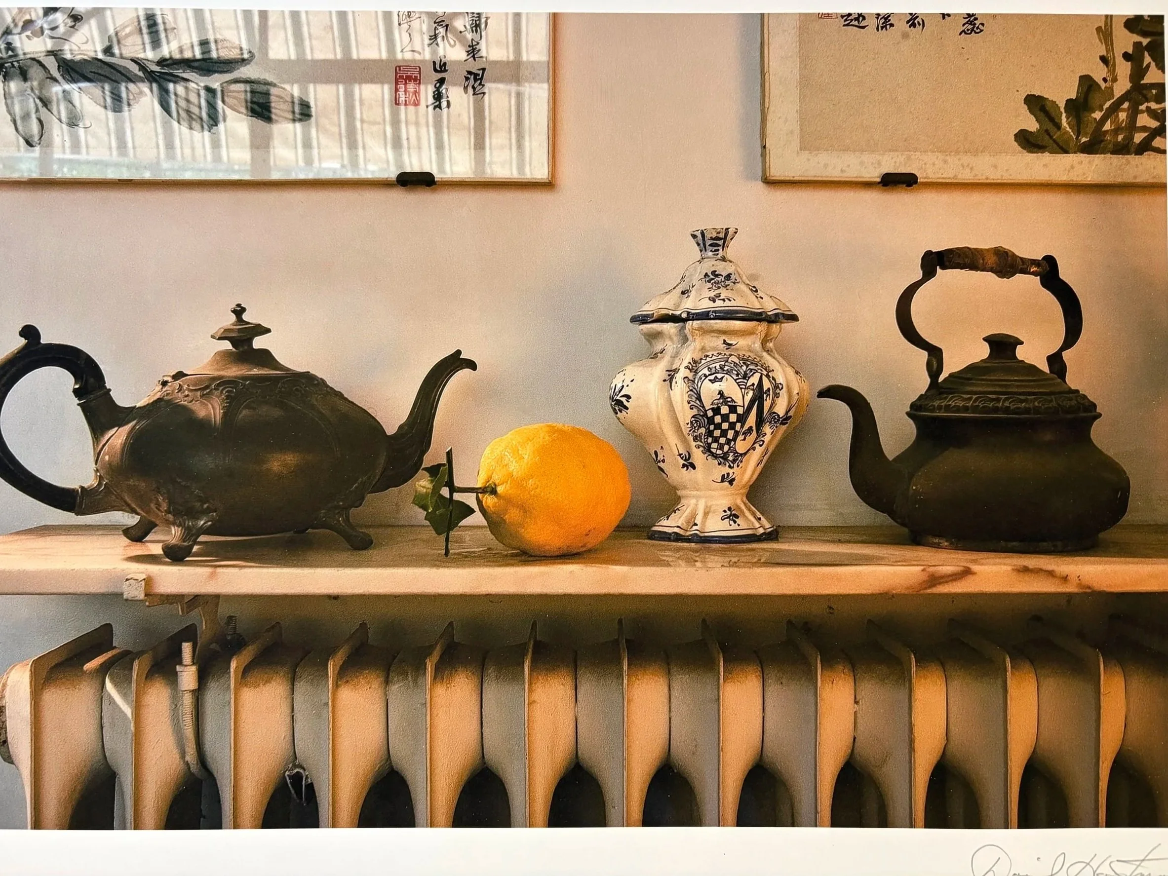 A display of vintage tea kettles and a decorative vase on a wooden shelf, with two Asian-style scroll paintings hanging on the wall above.