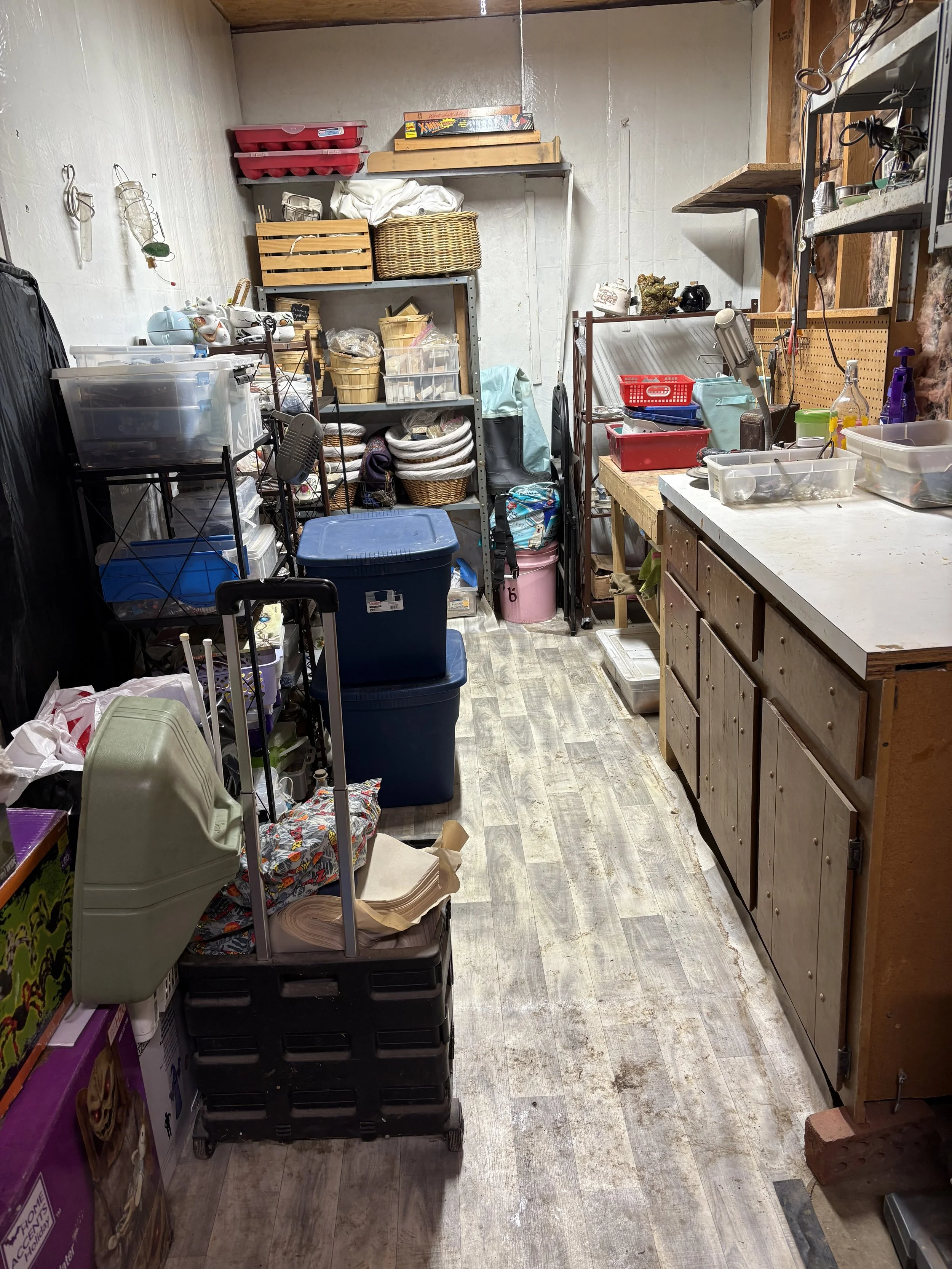 A cluttered storage or utility room with shelves filled with various household items and containers, a workbench with tools and cleaning supplies, and wooden flooring.