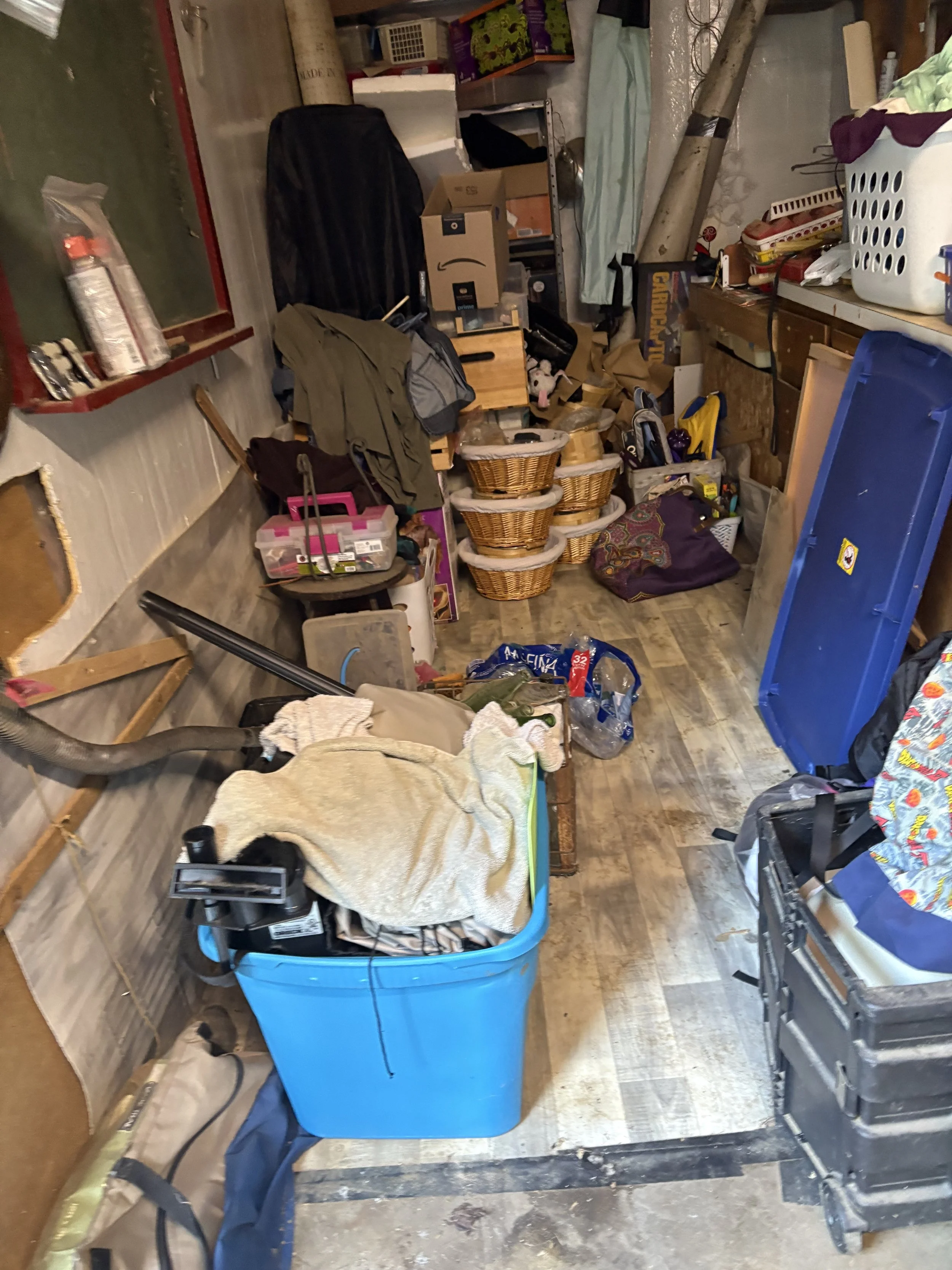 A cluttered room with various household items, including a large blue plastic bin, baskets, boxes, a vacuum cleaner, and miscellaneous objects on shelves and the floor, indicating storage or organization chaos.