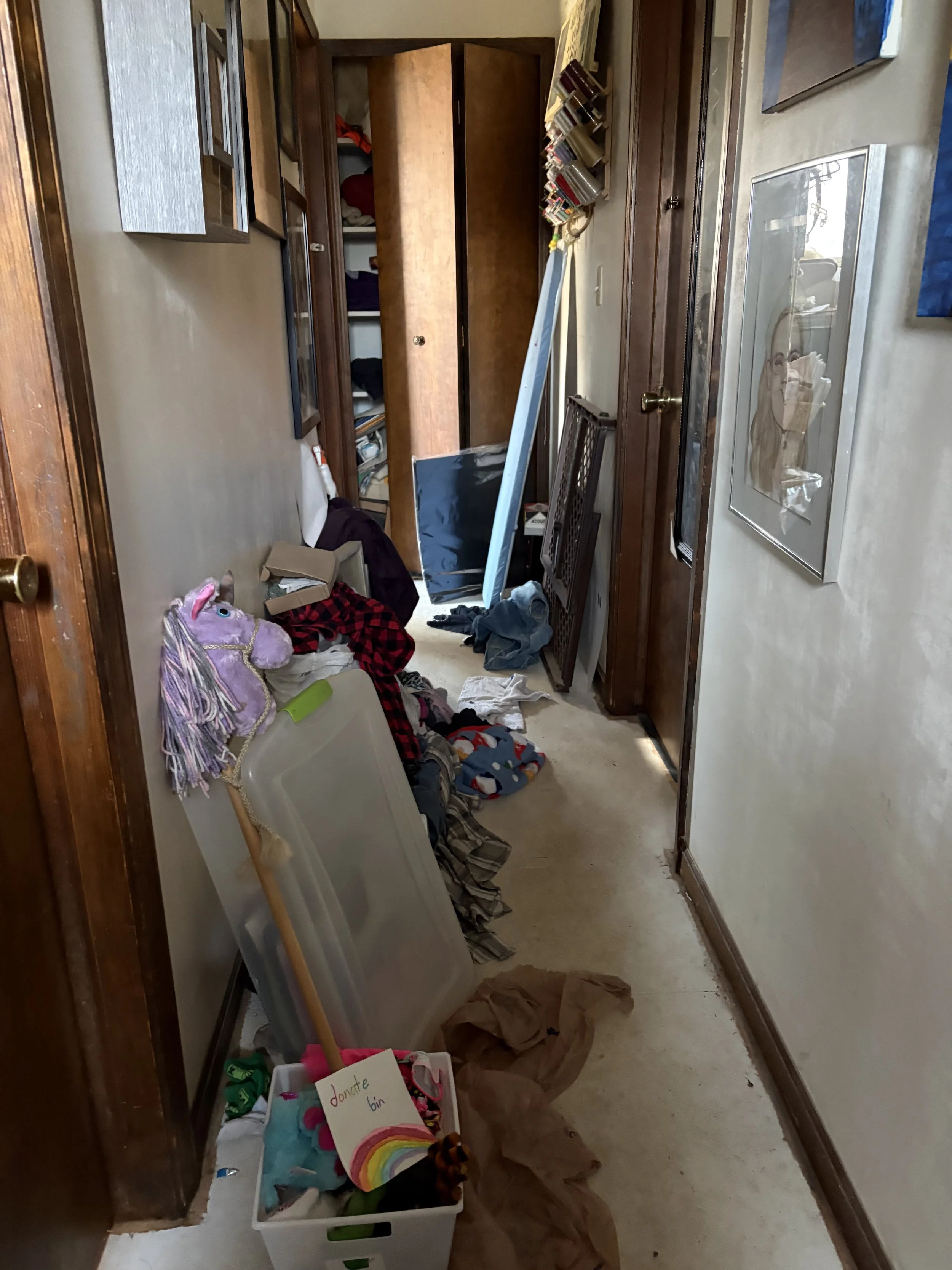 Cluttered hallway with open closet, scattered clothes, a plastic bin filled with toys and books, and a brown paper bag on the floor.