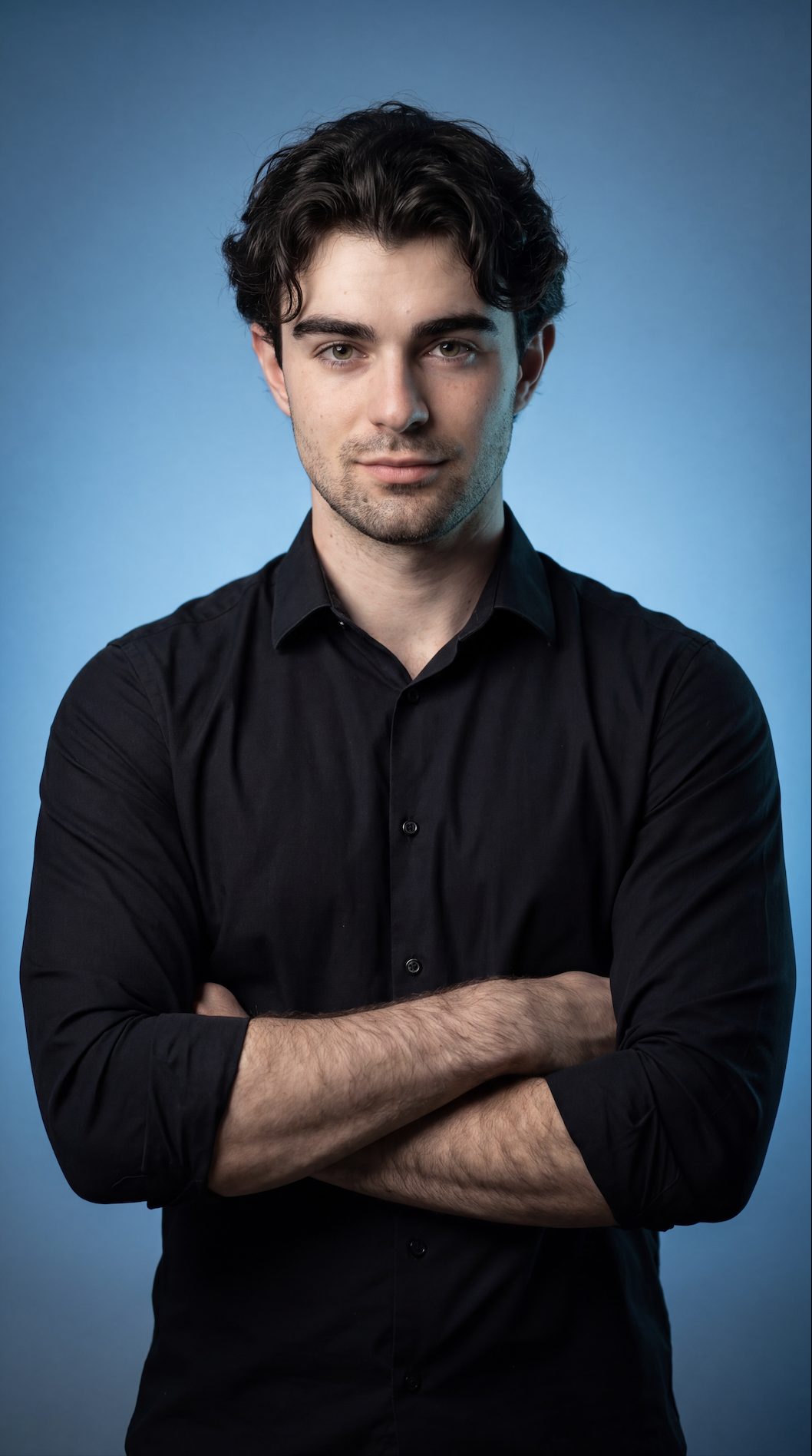 Portrait of a young man with dark curly hair, wearing a black button-up shirt, standing against a blue background with arms crossed.