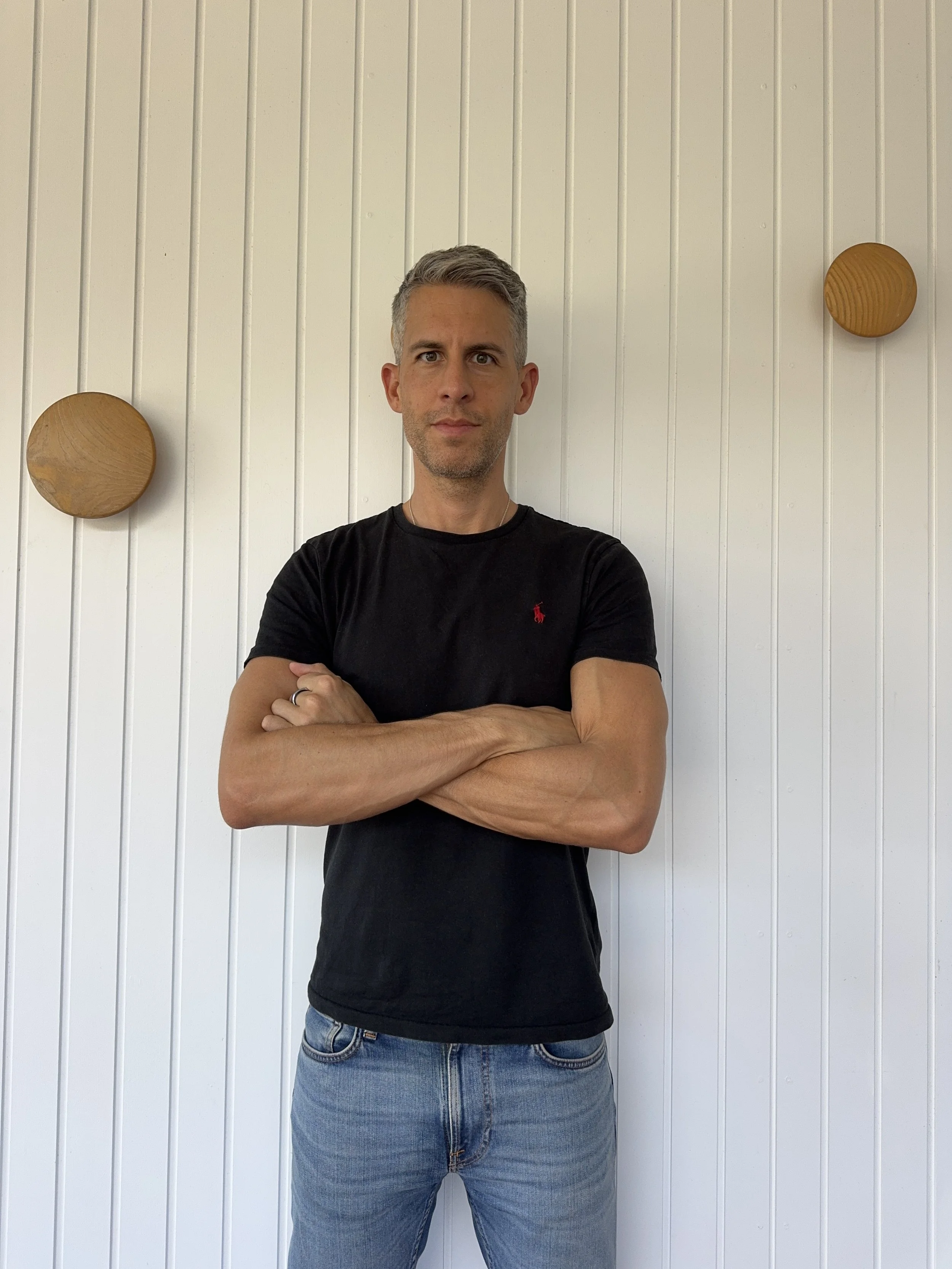 A man with gray hair and light skin posing with arms crossed in front of a white paneled wall with two round wooden decorations.