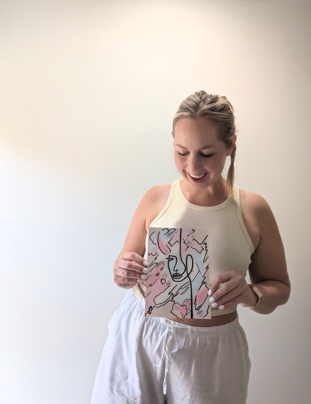 A woman with blonde hair styled in a braid, smiling and looking down at a piece of artwork she is holding. She is wearing a cream-colored sleeveless top and white drawstring pants, standing against a plain white wall.