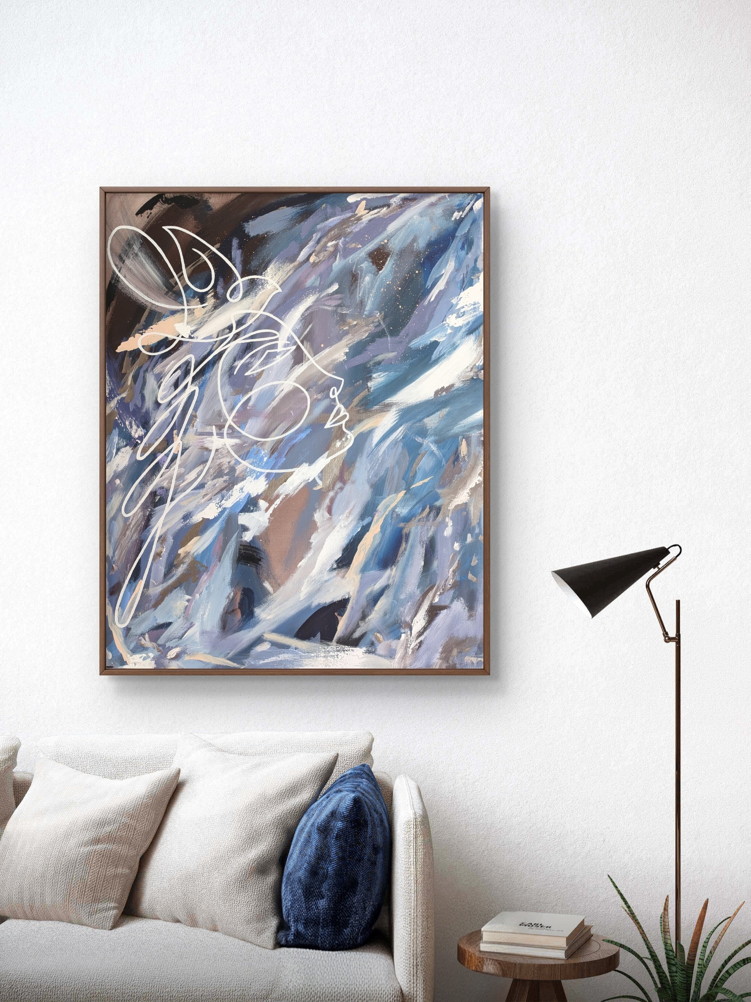 A living room corner with a beige sofa, blue patterned pillow, and an abstract painting with blue, white, black, and beige brushstrokes on the wall. A black floor lamp and a small side table with books are also visible.