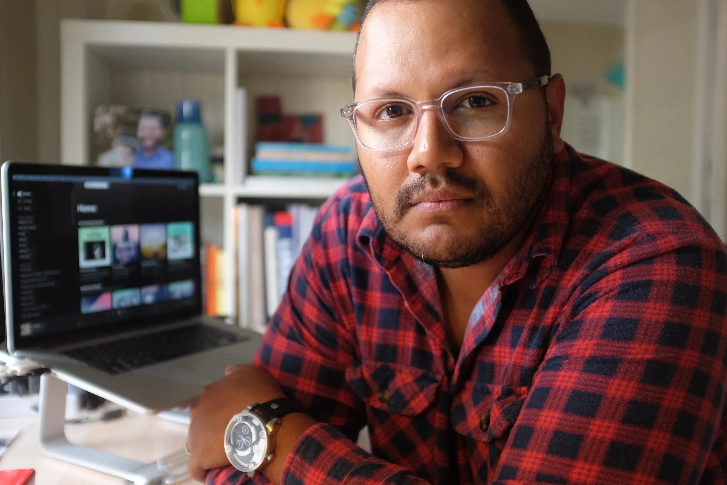 A man with short dark hair, glasses, and a beard wearing a red and black plaid shirt, sitting at a desk with a laptop, in a room with shelves filled with books, colorful bowls, and a small framed photo in the background.