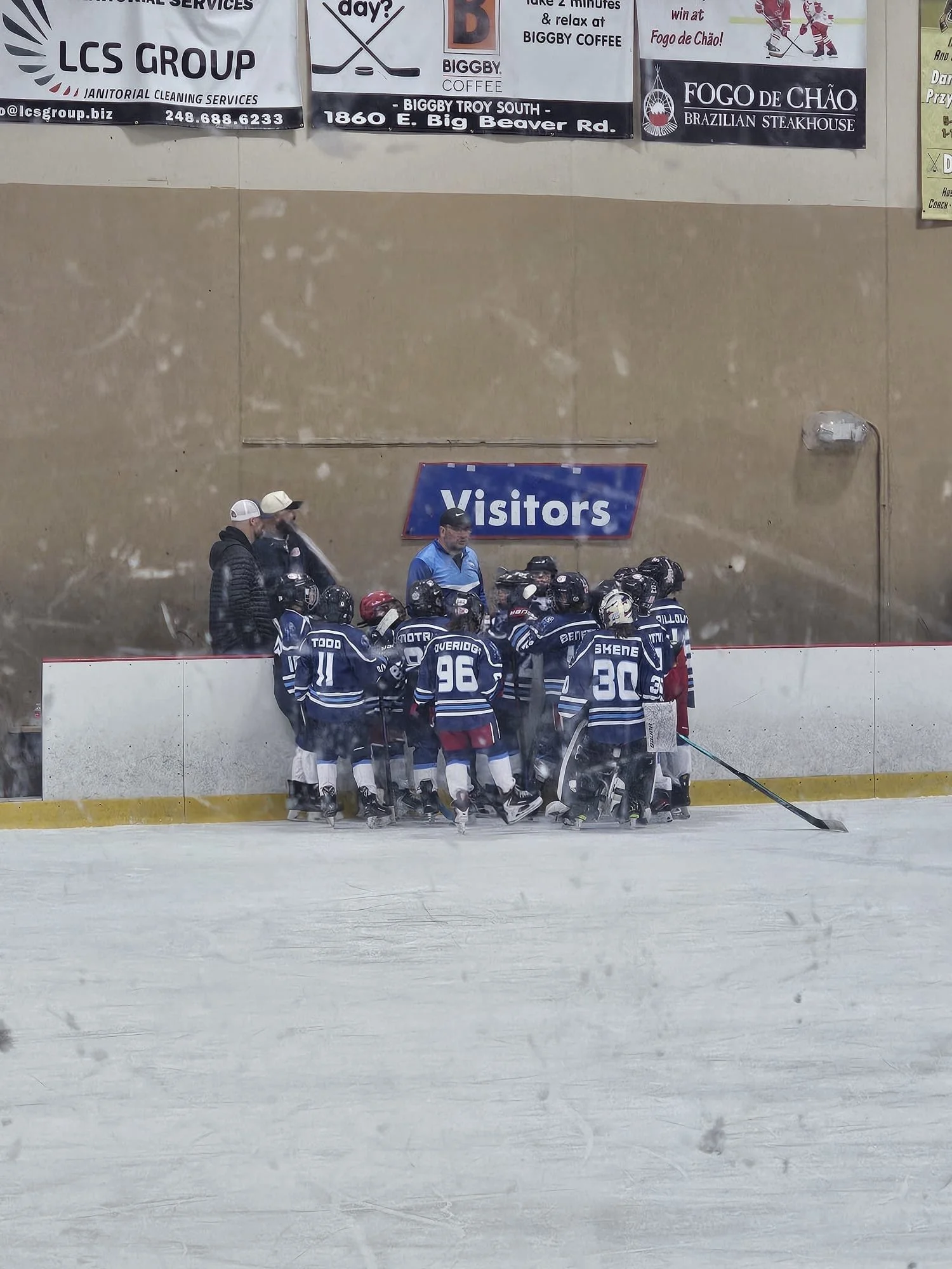 Hockey team in dark blue jerseys gathered near the side of an ice rink, meeting with their coaches or trainers. A sign labeled 'Visitors' is visible behind them.