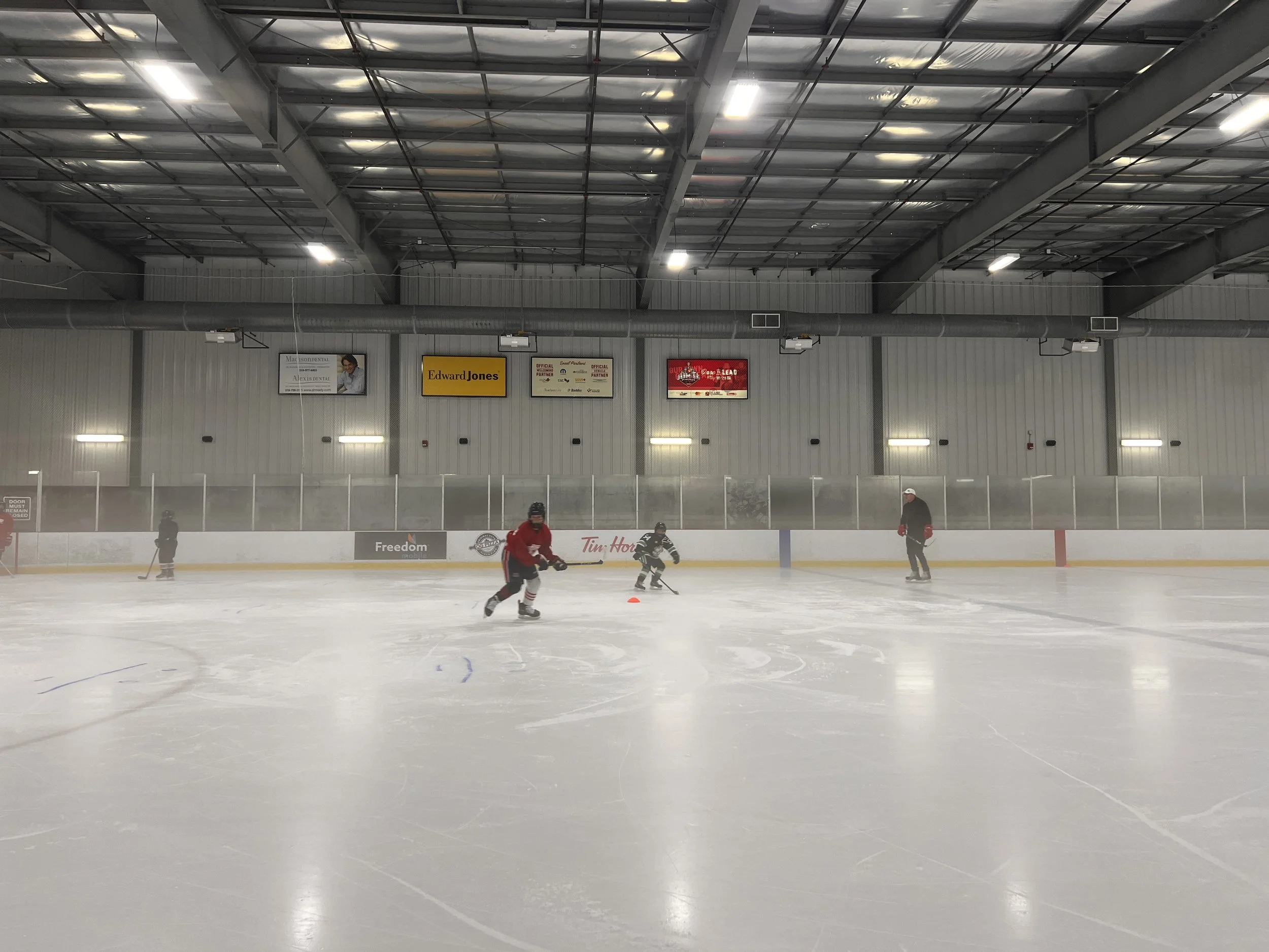 Ice hockey rink with players skating on the ice and a person skating in the background, with advertising banners hanging on the wall above the rink.