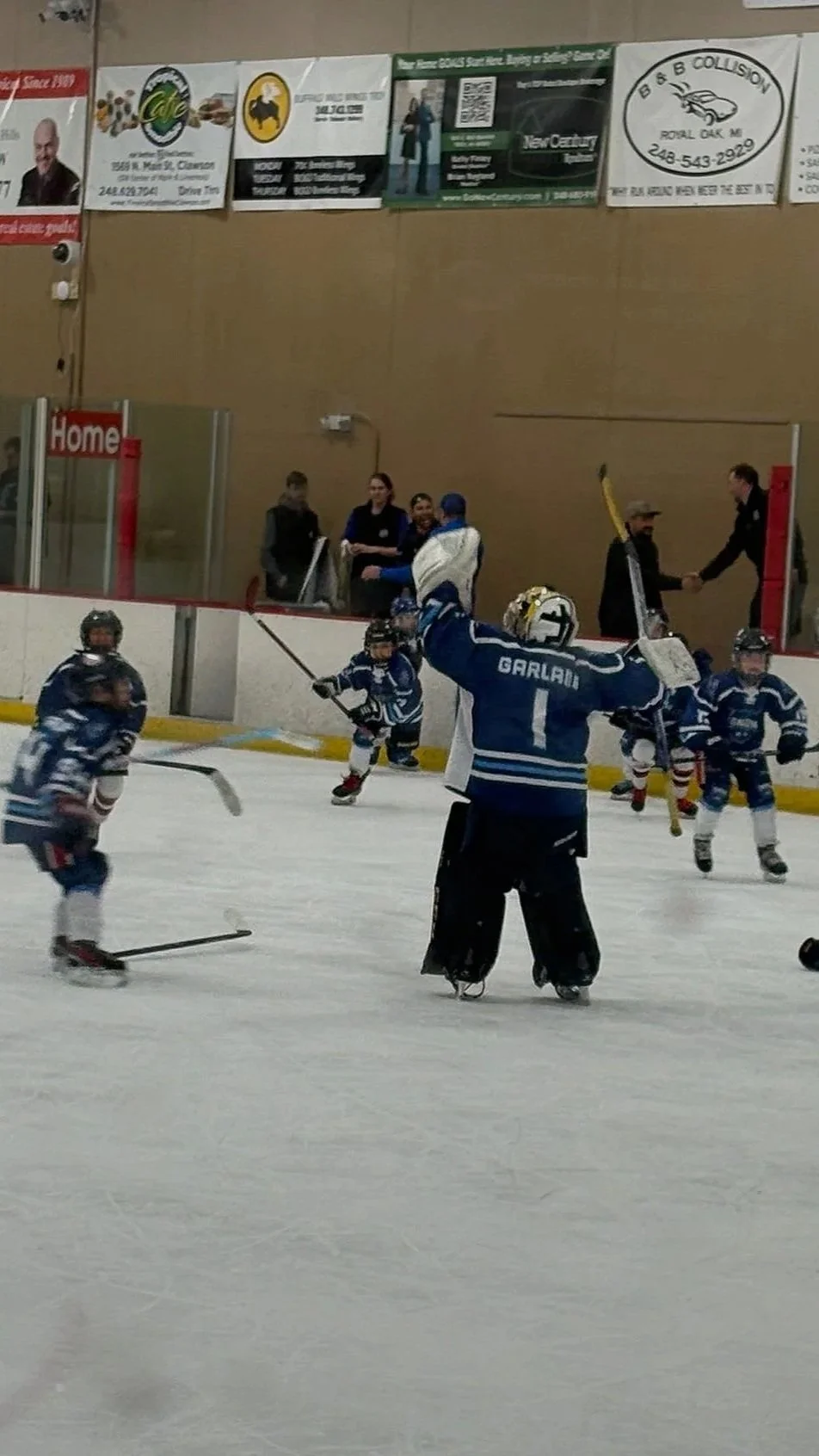 A hockey rink with young children playing hockey, led by a coach wearing a jersey with the name 'Garlan' and the number 1. The coach is celebrating with arms raised. Spectators and officials are visible in the background near the glass wall.