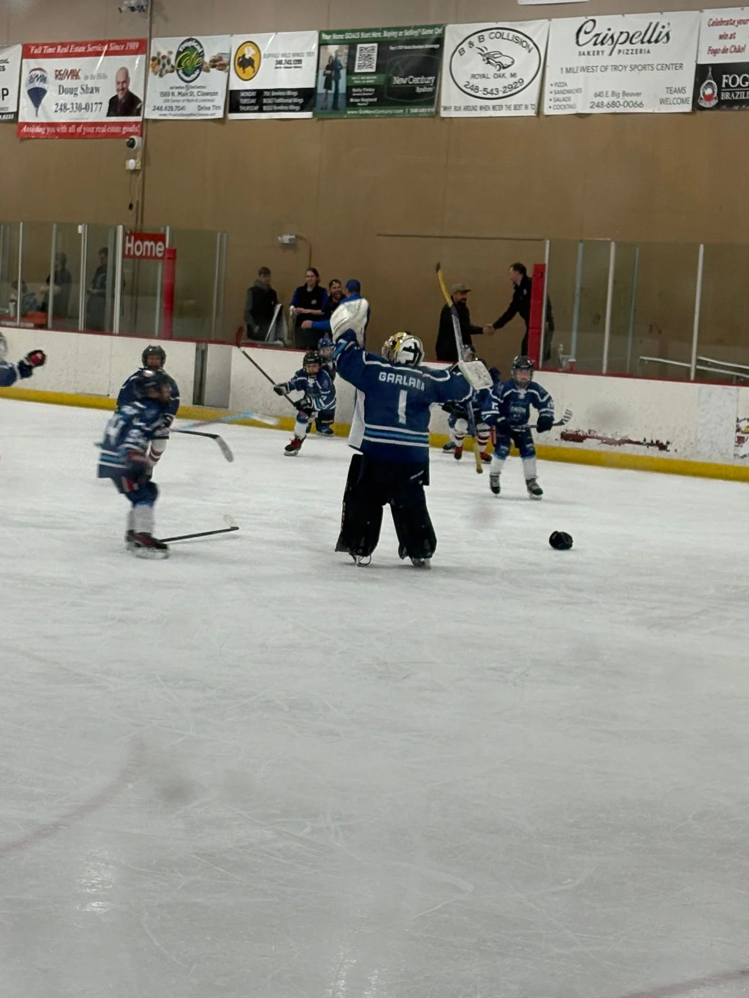 A hockey game in progress at an indoor ice rink. A goalie in blue is celebrating with a raised stick, while several players and a referee are on the ice. Spectators and coaches watch from behind the glass.