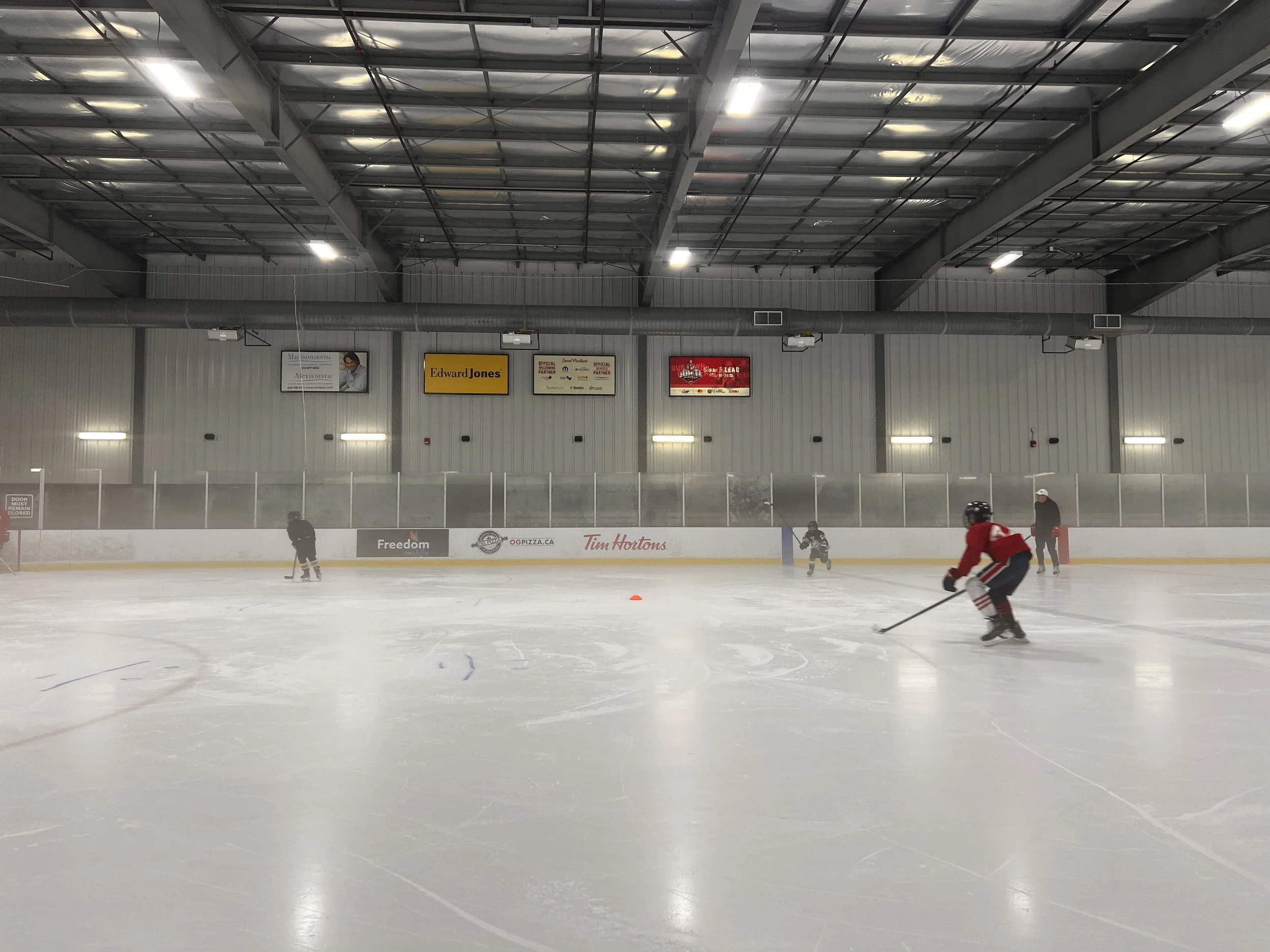 Indoor ice hockey rink with players practicing skating and handling pucks, banners on the wall, and a figure skating in the background.