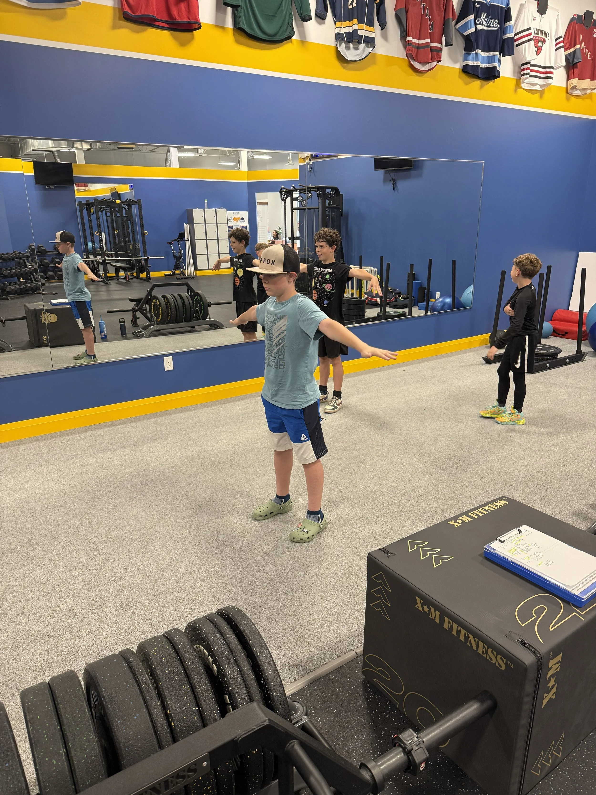 Children in a gym studio standing on an exercise mat, stretching with arms outstretched. The gym has a large mirror, fitness equipment, weights, and a box labeled 'X-M FITNESS'.