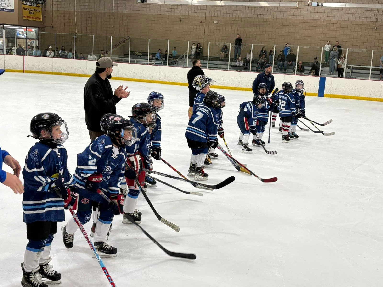 A group of young hockey players in blue jerseys and helmets lined up on the ice rink, with coaches standing nearby, during a game or practice session at an indoor ice hockey arena.