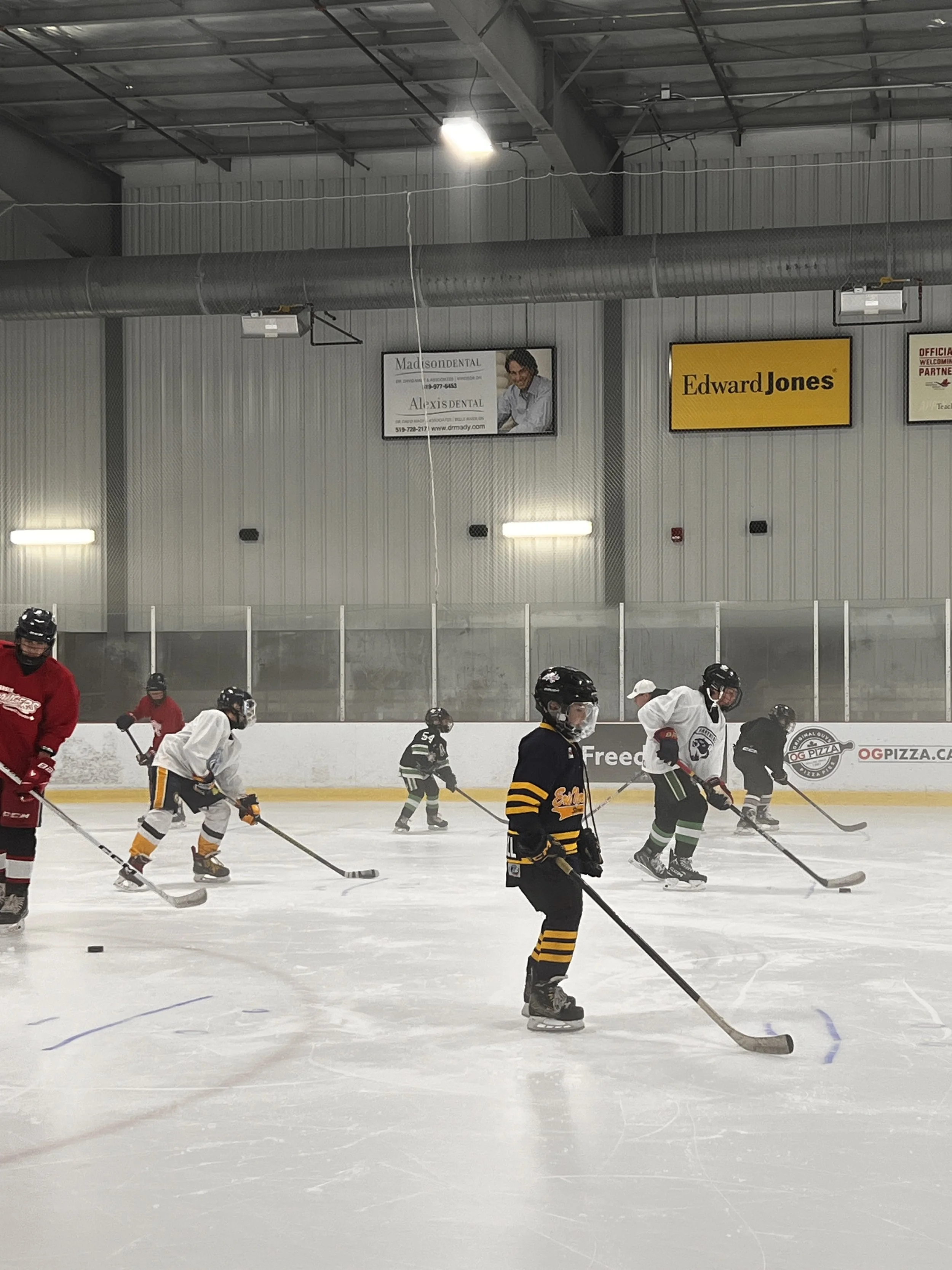 Young players practicing hockey on an indoor ice rink, coached by adults, wearing helmets and jerseys, with a wall featuring advertising banners in the background.
