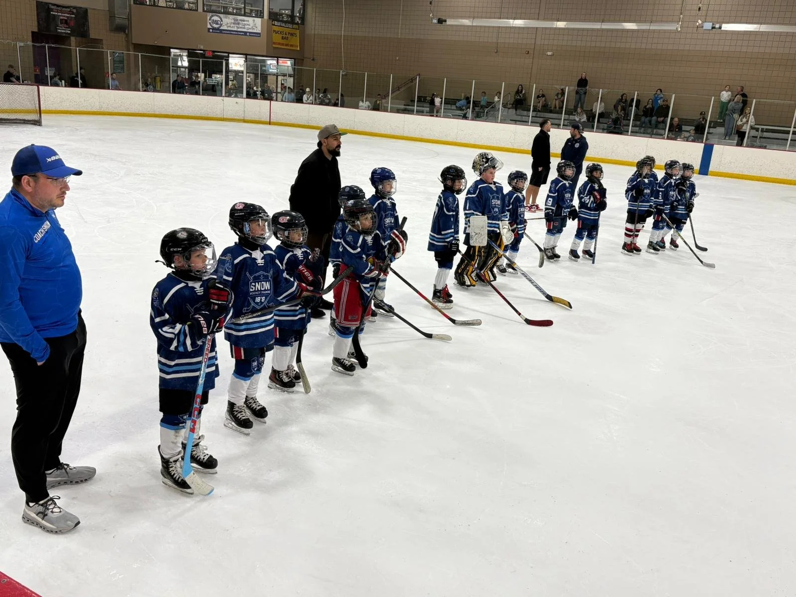 A group of young hockey players in blue uniforms with helmets and sticks standing on an ice rink, lined up for a lineup with coaches and spectators in the background.