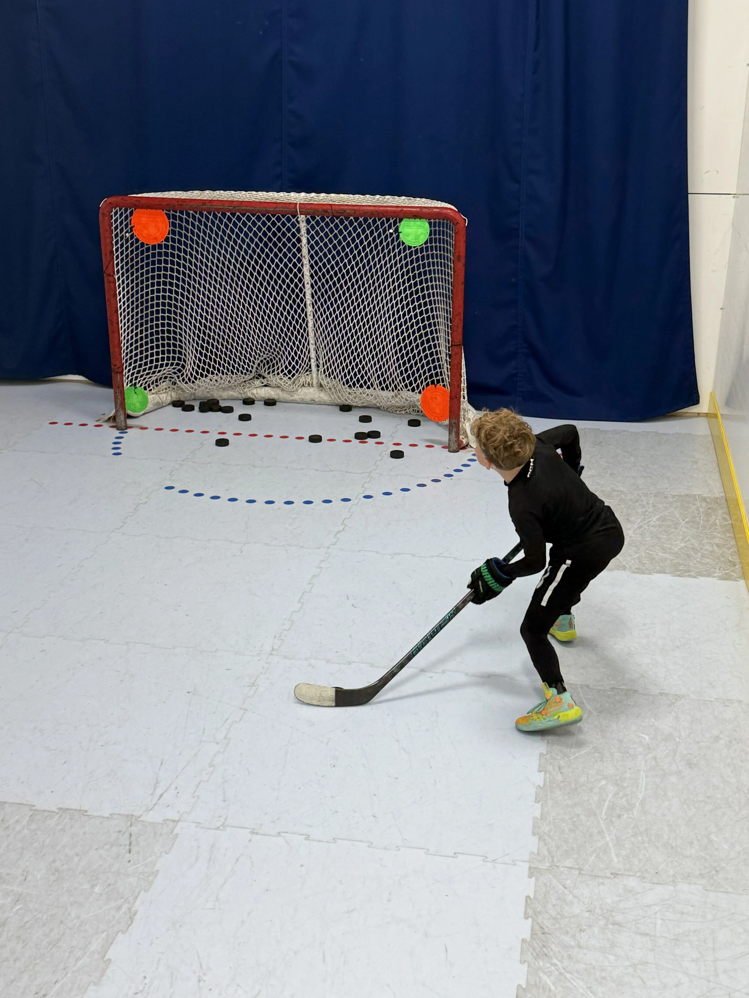 A young boy wearing colorful sneakers playing hockey indoors. The boy is bent over with a hockey stick, in front of a hockey net with orange and green balls attached to the top of the net. The floor has lines and dots, and a blue curtain is in the background.