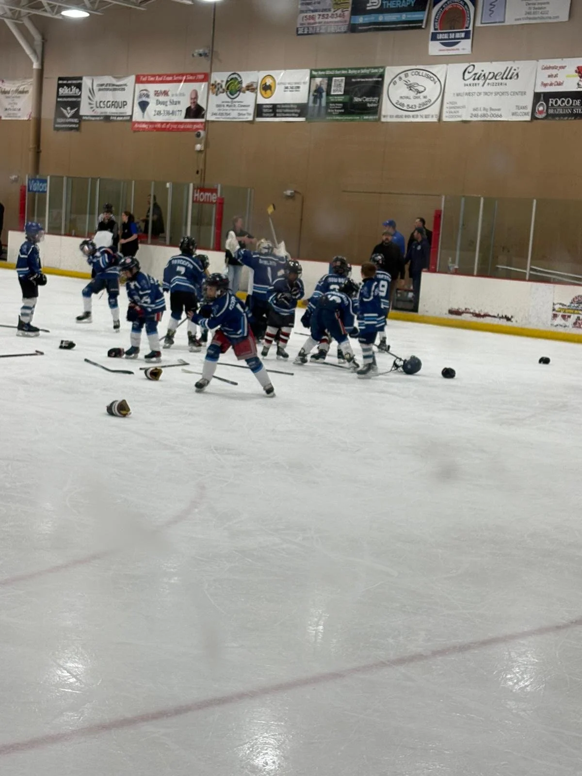 Children in hockey uniforms practicing or playing on an ice rink, with hockey sticks and helmets scattered around, and a few adults observing nearby.
