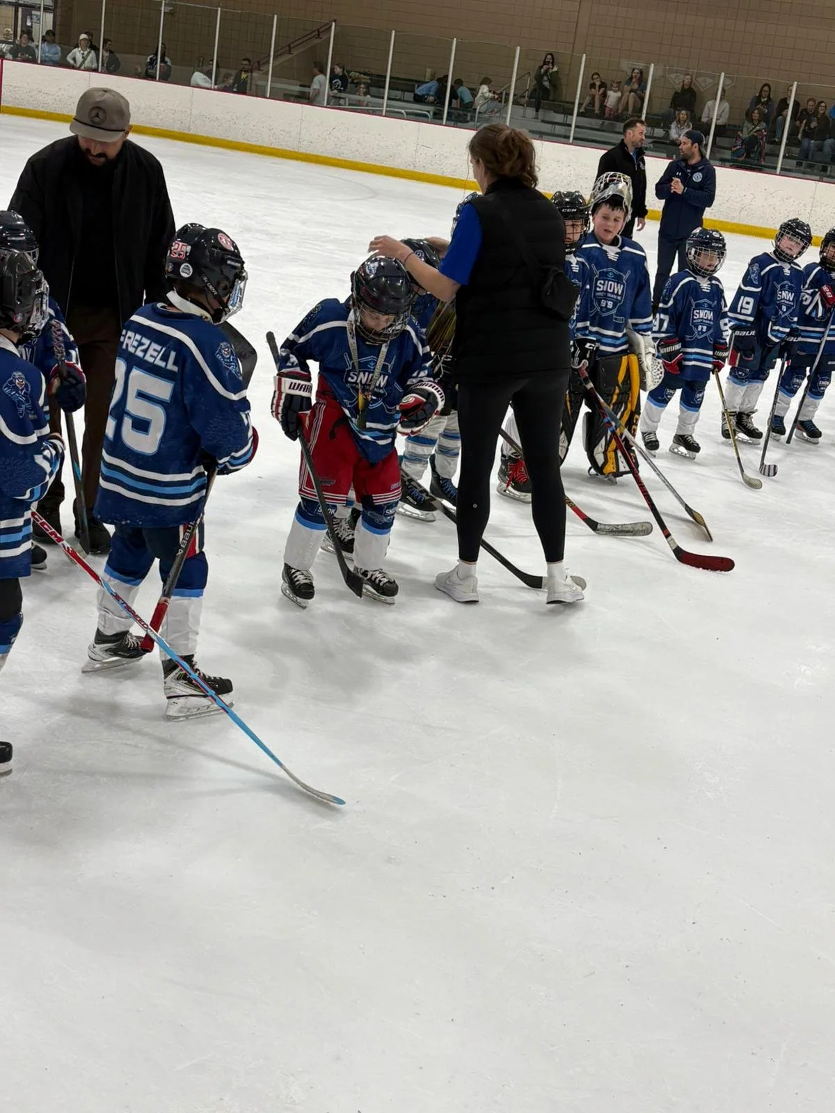 Children in hockey uniforms standing in line on ice rink, with coaches and audience watching in background.