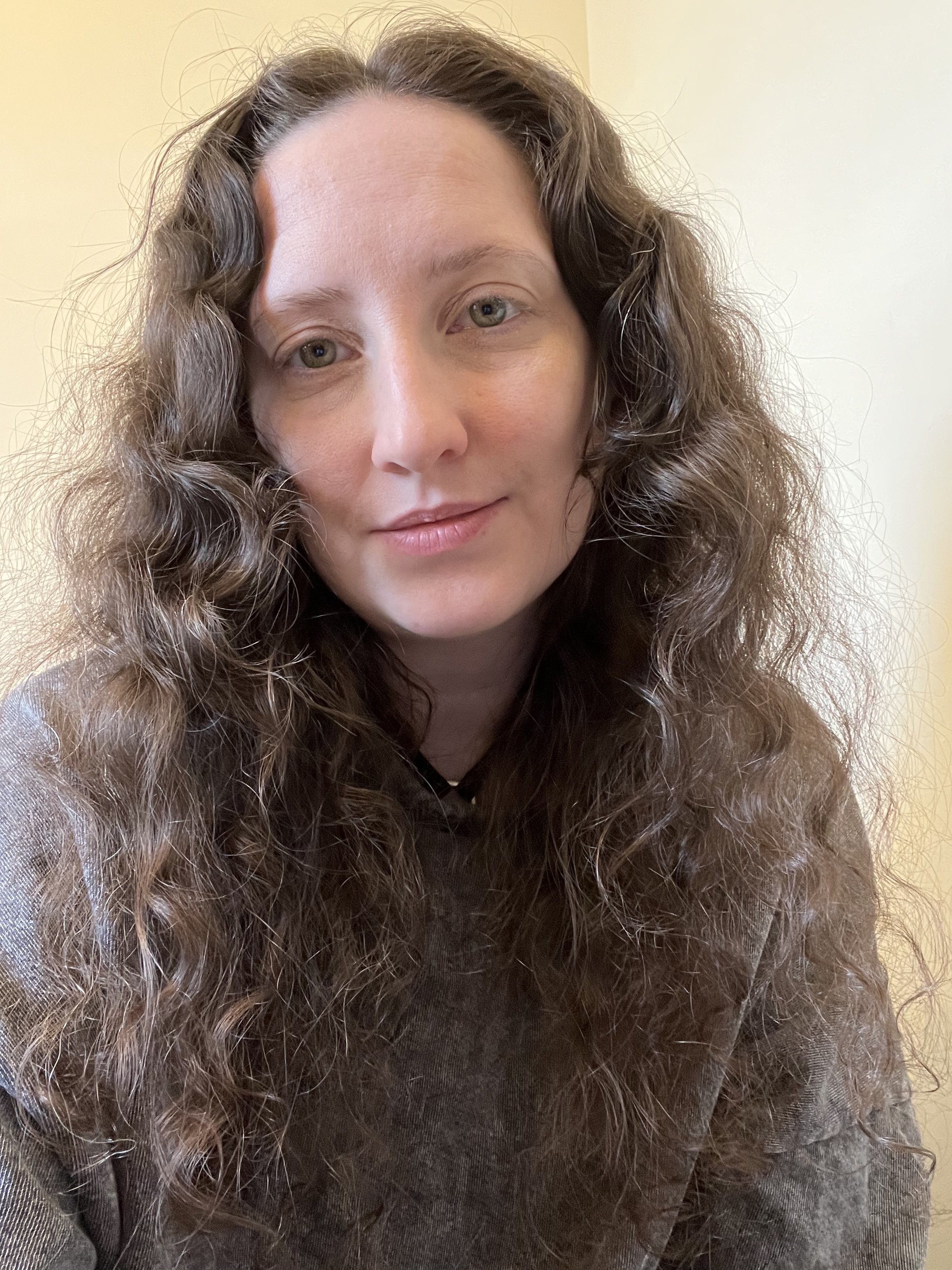 A woman with long, curly brown hair and light skin looking directly at the camera with a neutral expression, wearing a dark top and standing in front of a beige wall.