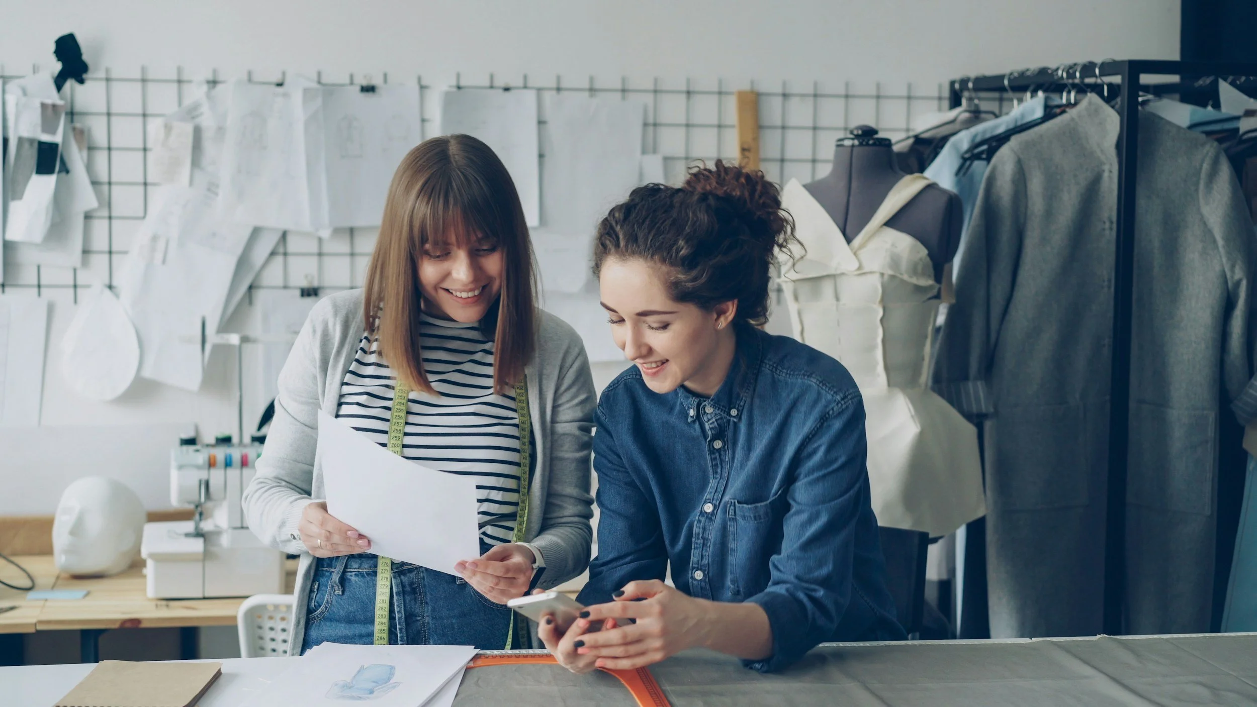 Two women in a design studio looking at a phone and papers, with sewing patterns and mannequins in the background.