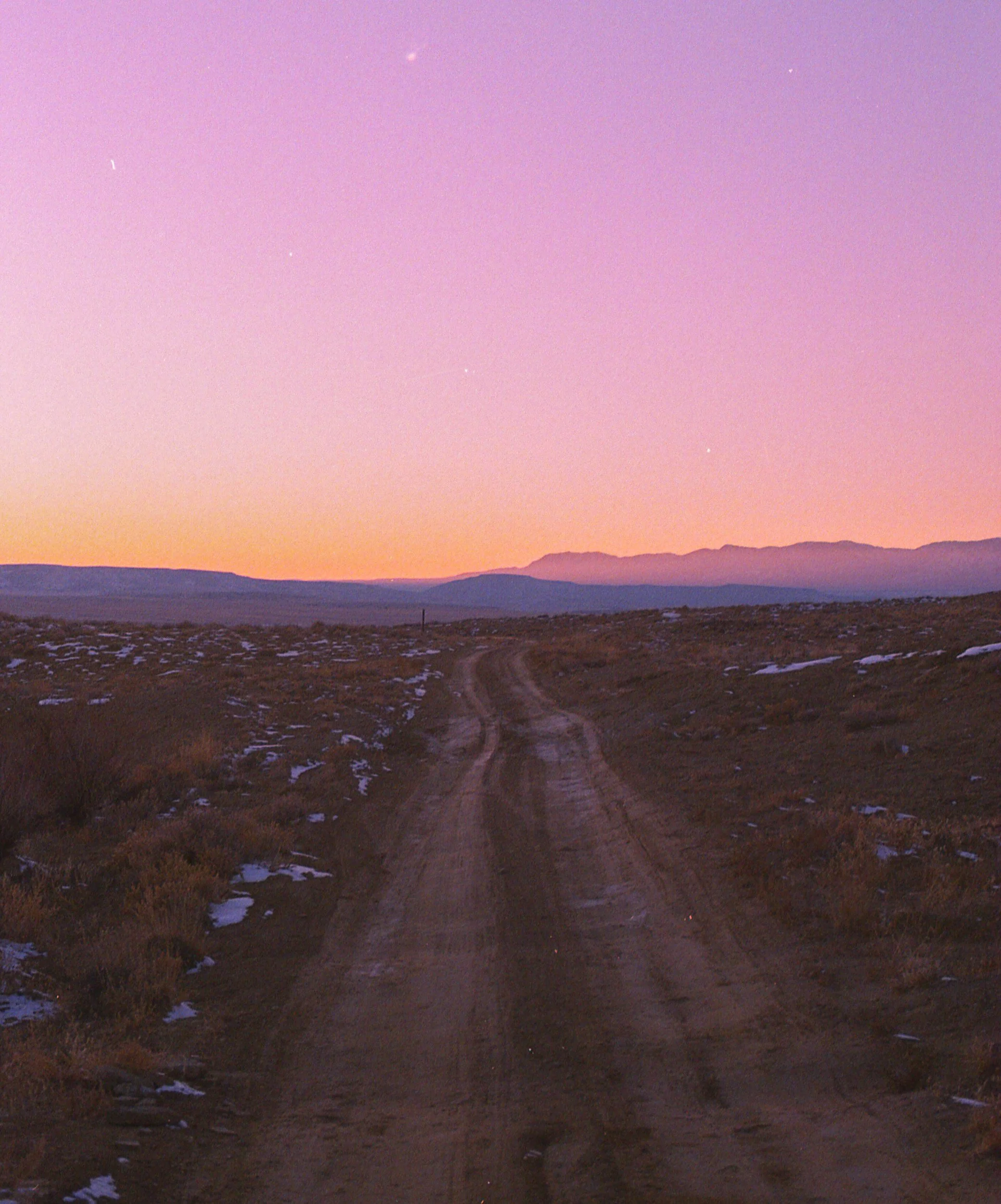 Dirt road running through a landscape with sparse vegetation, patches of snow, mountains in the distance, and a pink and purple sunset sky.
