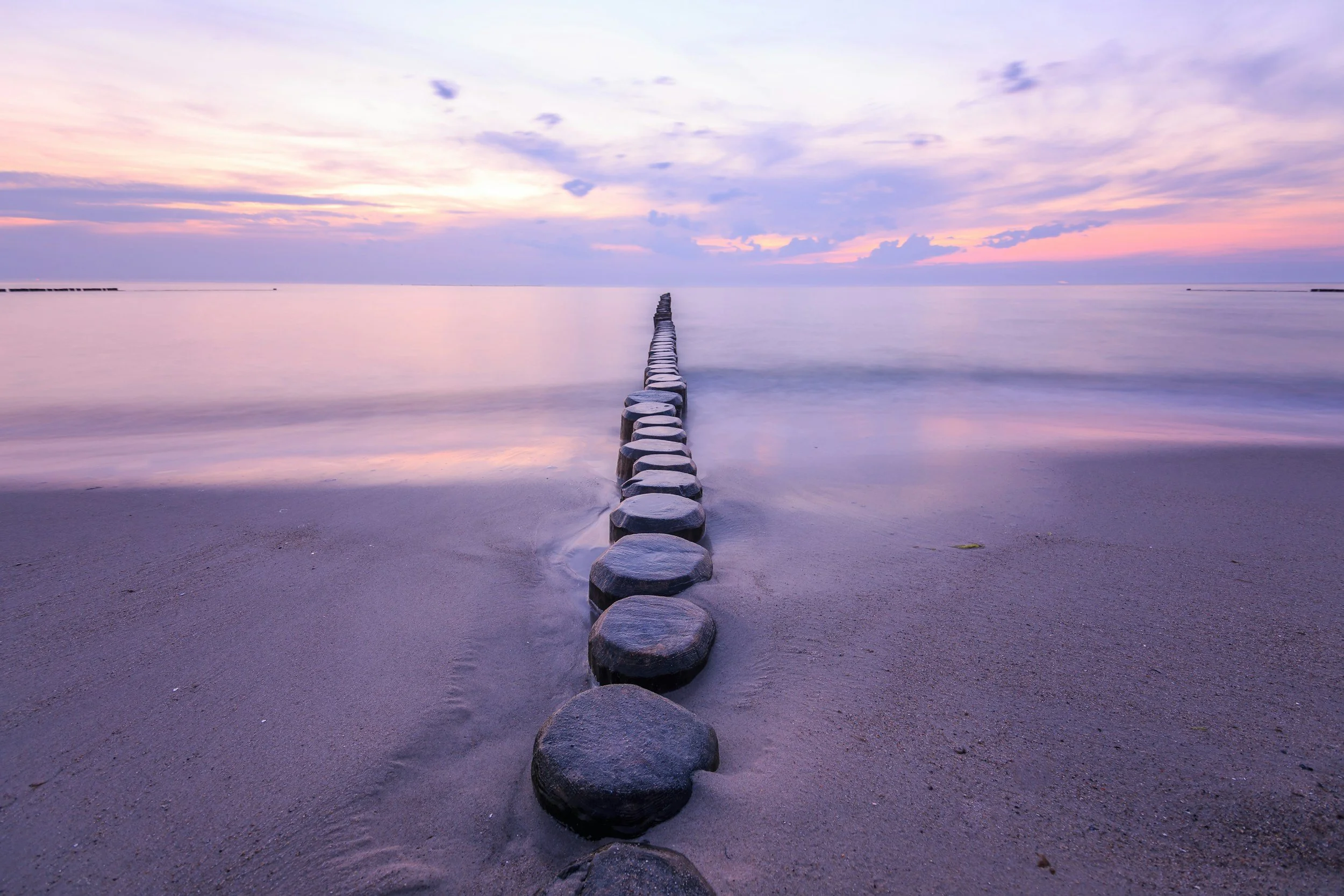 A serene beach scene at sunset with pastel purple and pink sky, calm water, and a line of smooth dark stones stretching into the horizon.