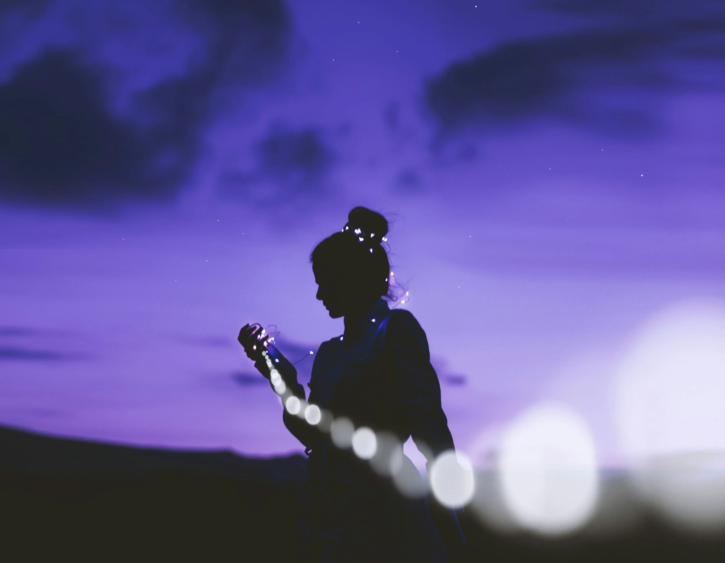 Silhouette of a woman with a bun hairstyle standing outdoors at night, holding a string of fairy lights that illuminate her outline, with a purple and blue sky and clouds in the background.