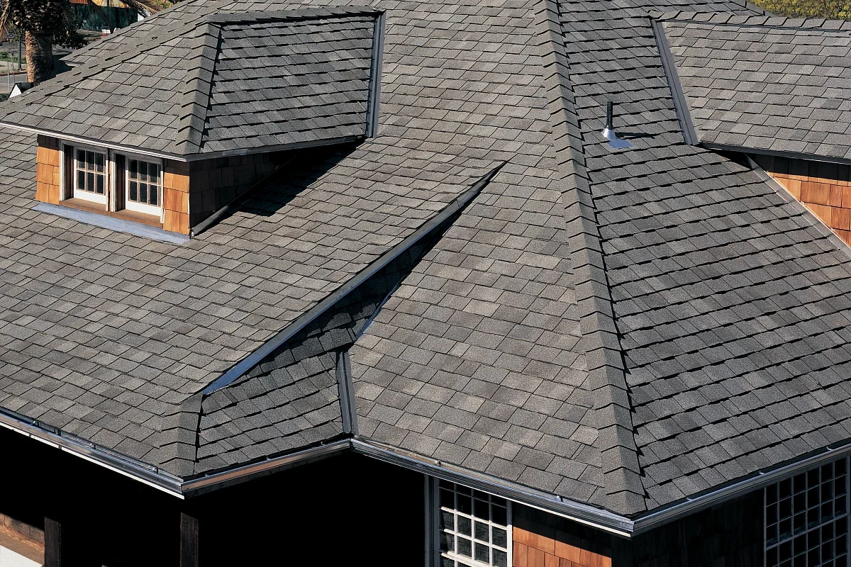 An aerial view of a house roof with gray asphalt shingles, featuring a dormer window and metal flashing along the edges.
