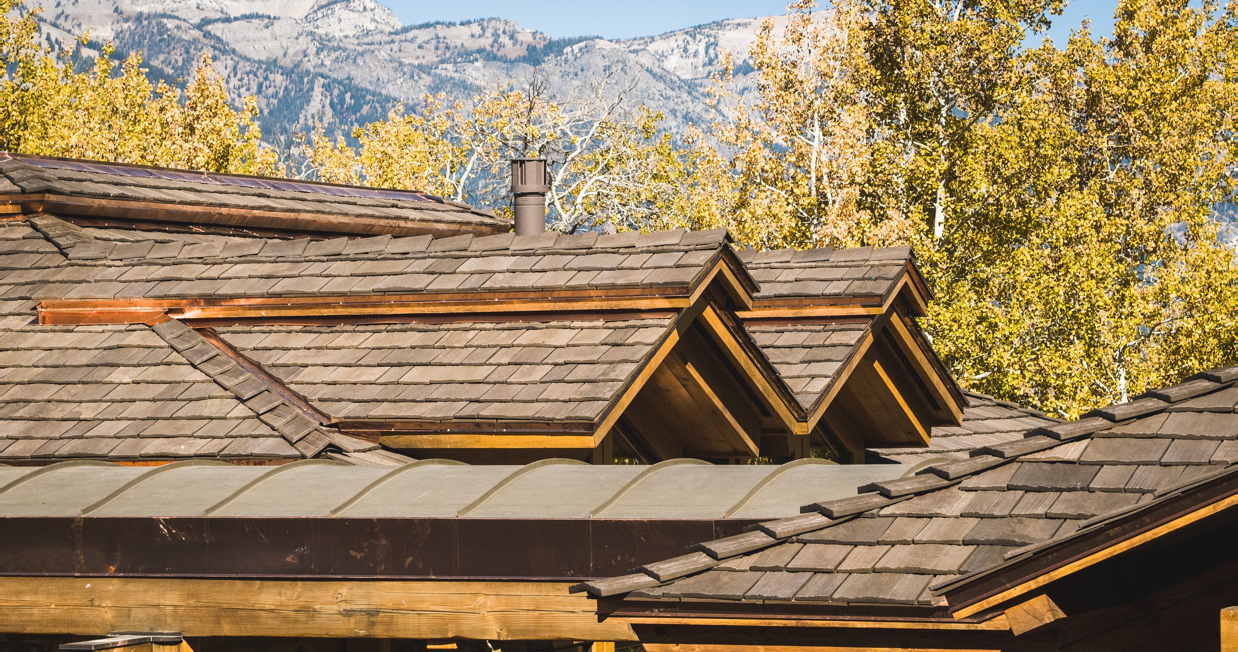 Close-up view of a wooden house roof with brown shingles, adjacent to a metal roof. The background shows autumn trees with yellow leaves and snow-capped mountains.