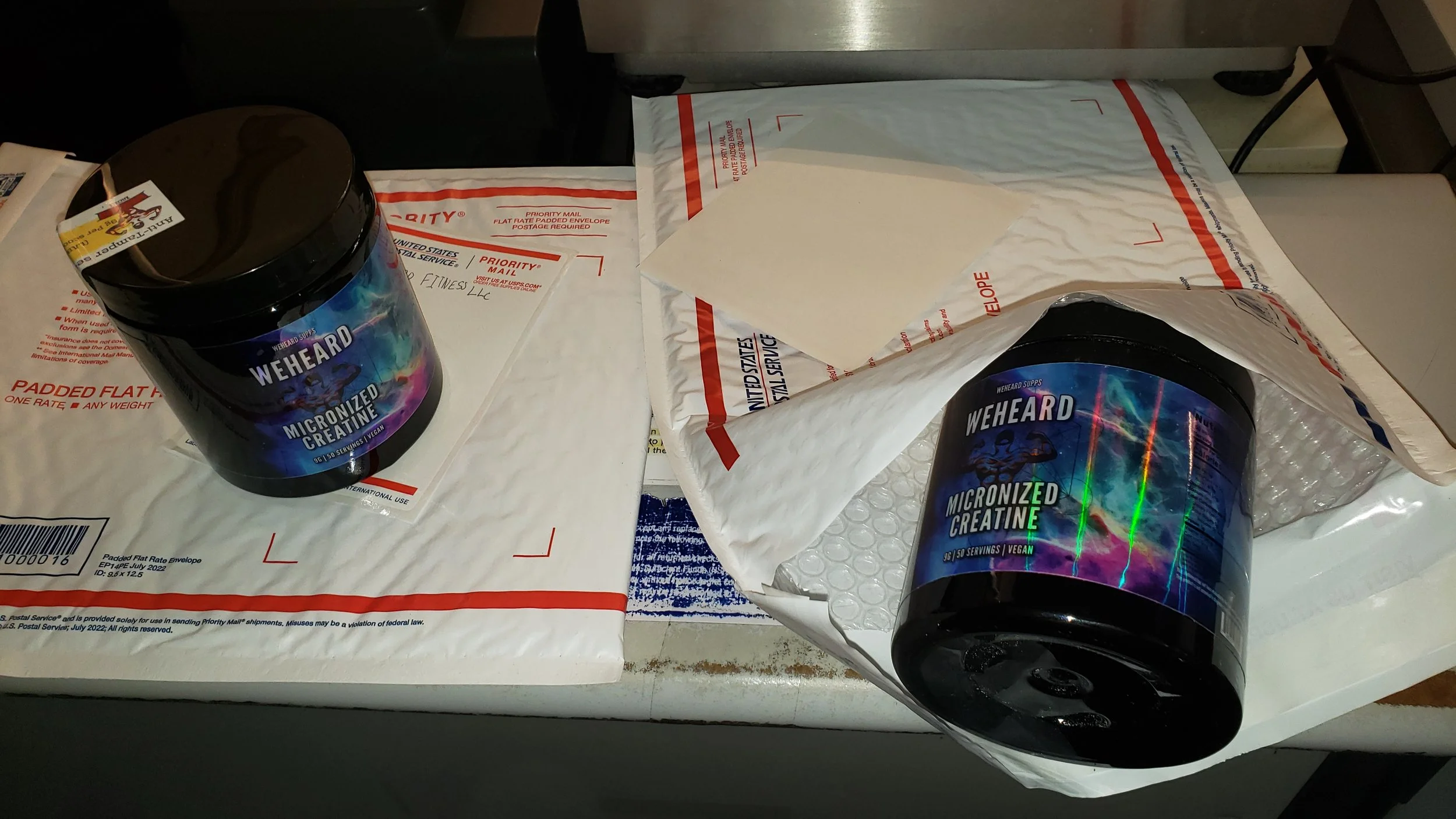 Two black containers of We Heard Micronized Creatine on a United States Postal Service padded flat-rate envelope, with one container on the envelope and the other partially inside a plastic bag, on a table.