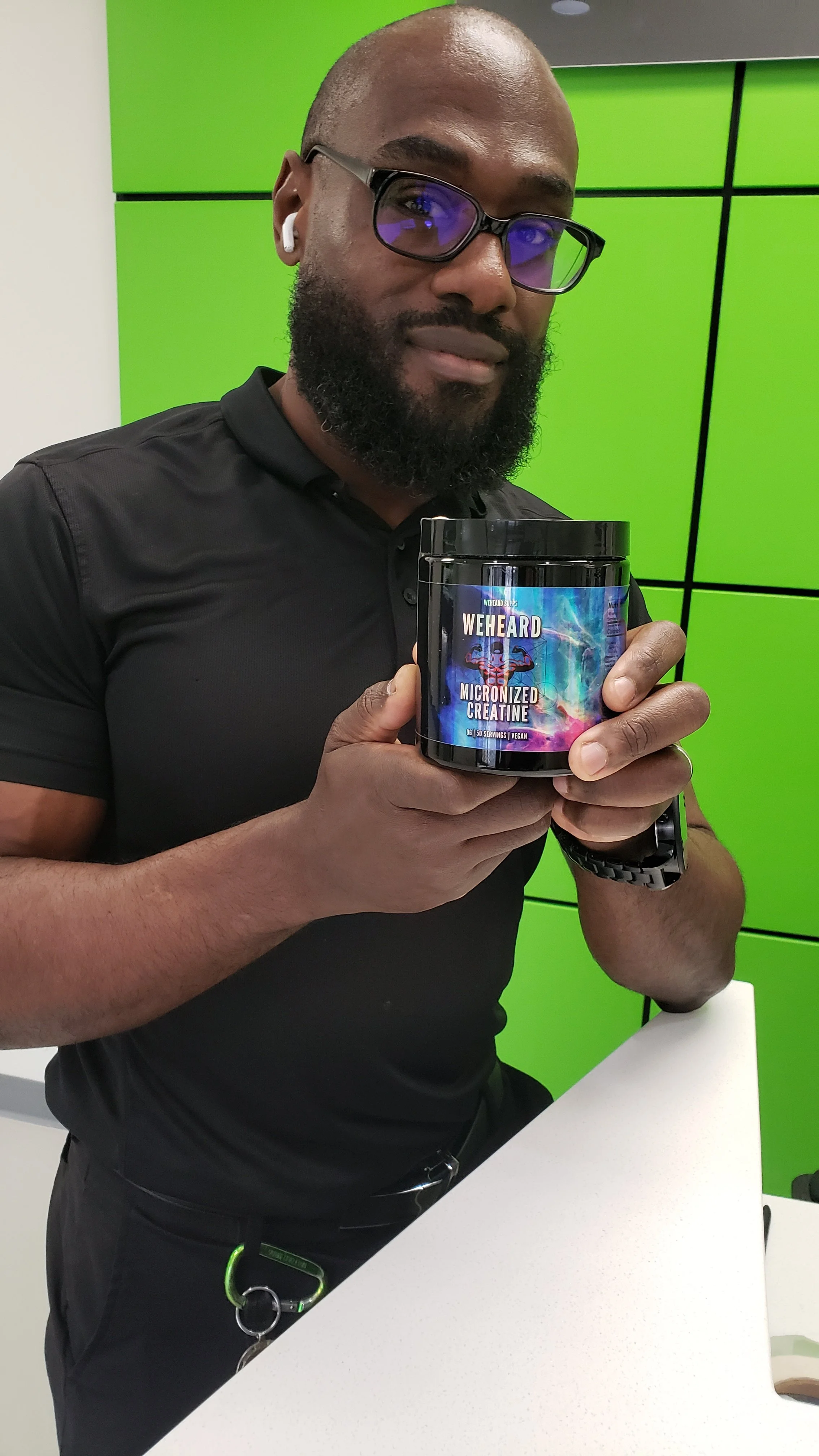A man with glasses, a beard, and earbuds holding a container labeled "WEHEARD MICRONIZED CREATINE" in a room with green and black geometric wall panels.