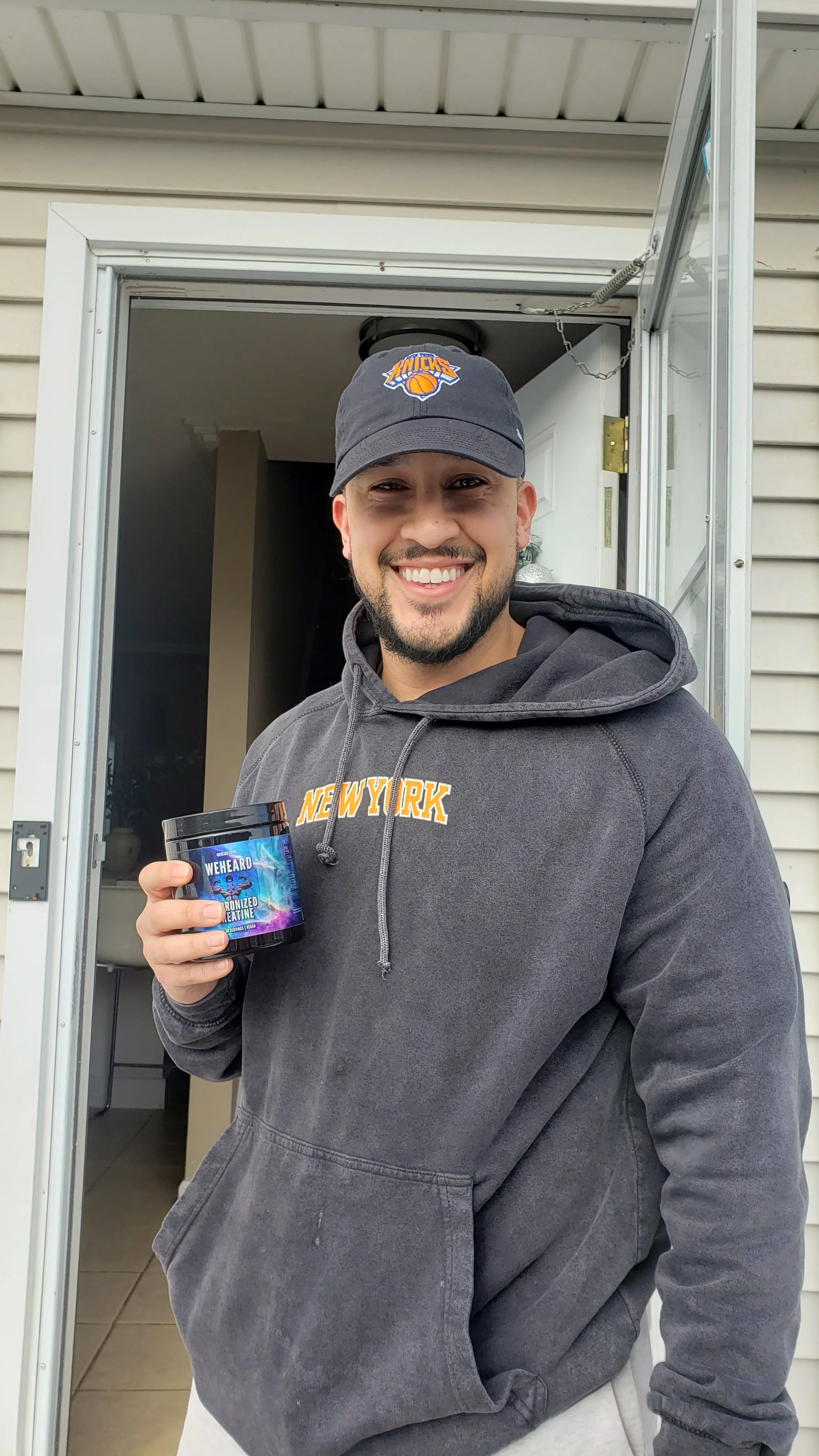 A smiling man standing at a doorway, wearing a New York hoodie and baseball cap, holding a container labeled 'WEHEARD' and 'MICRONIZED CRETINE'.