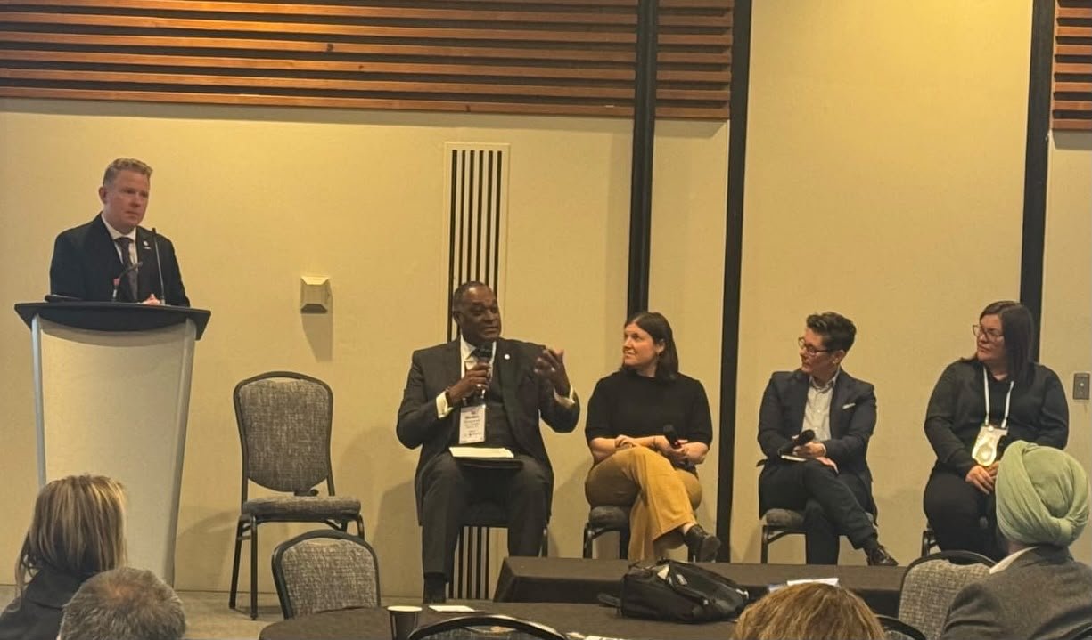 A panel discussion with five people, one standing at a podium and four seated, in a conference room.