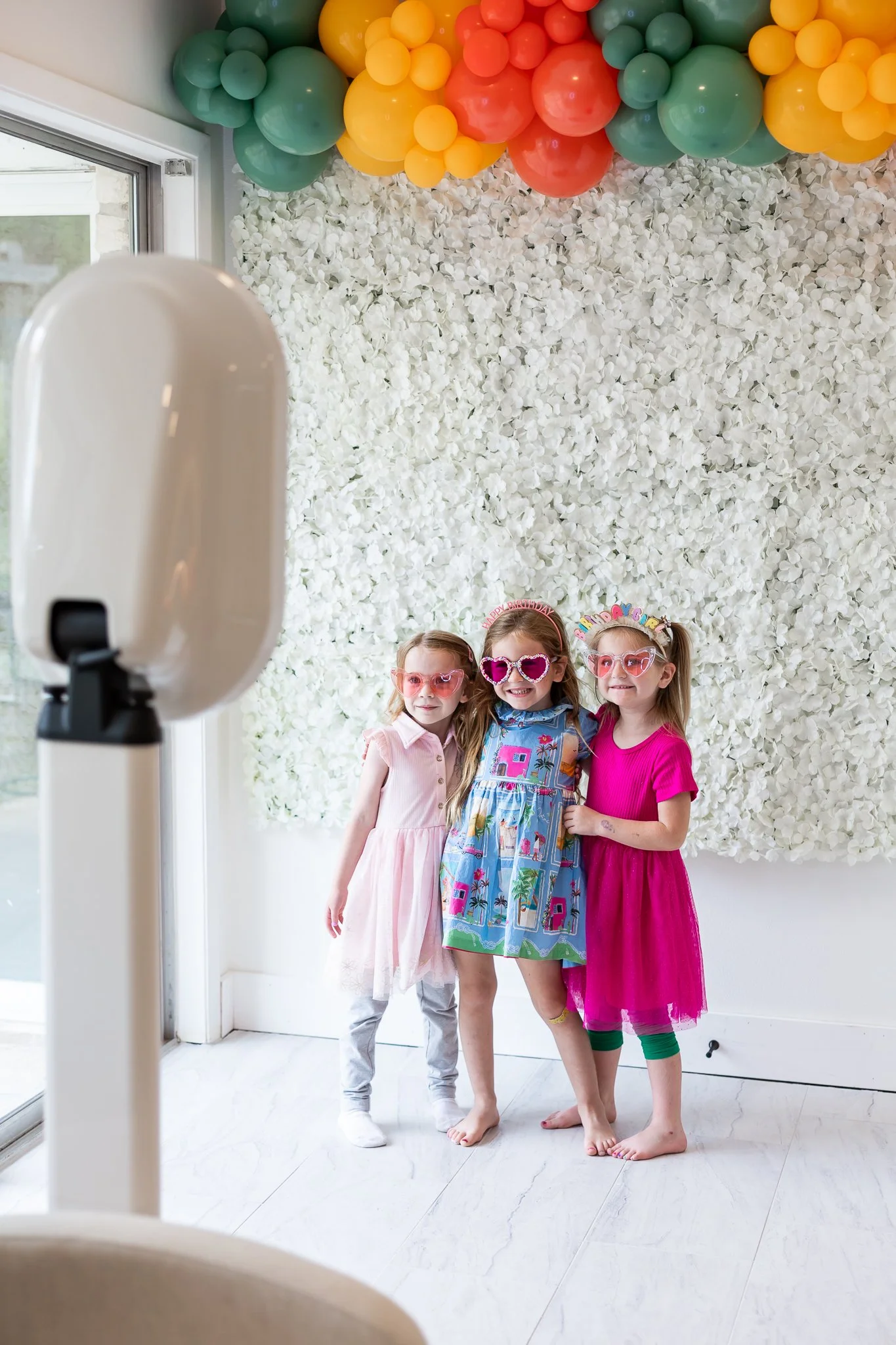 Three young girls posing for a photo at a birthday party, standing on a white marble floor against a background of white flower wall decor and a colorful balloon arch with green, yellow, and orange balloons. All three girls are wearing colorful dresses and playful glasses, smiling happily.