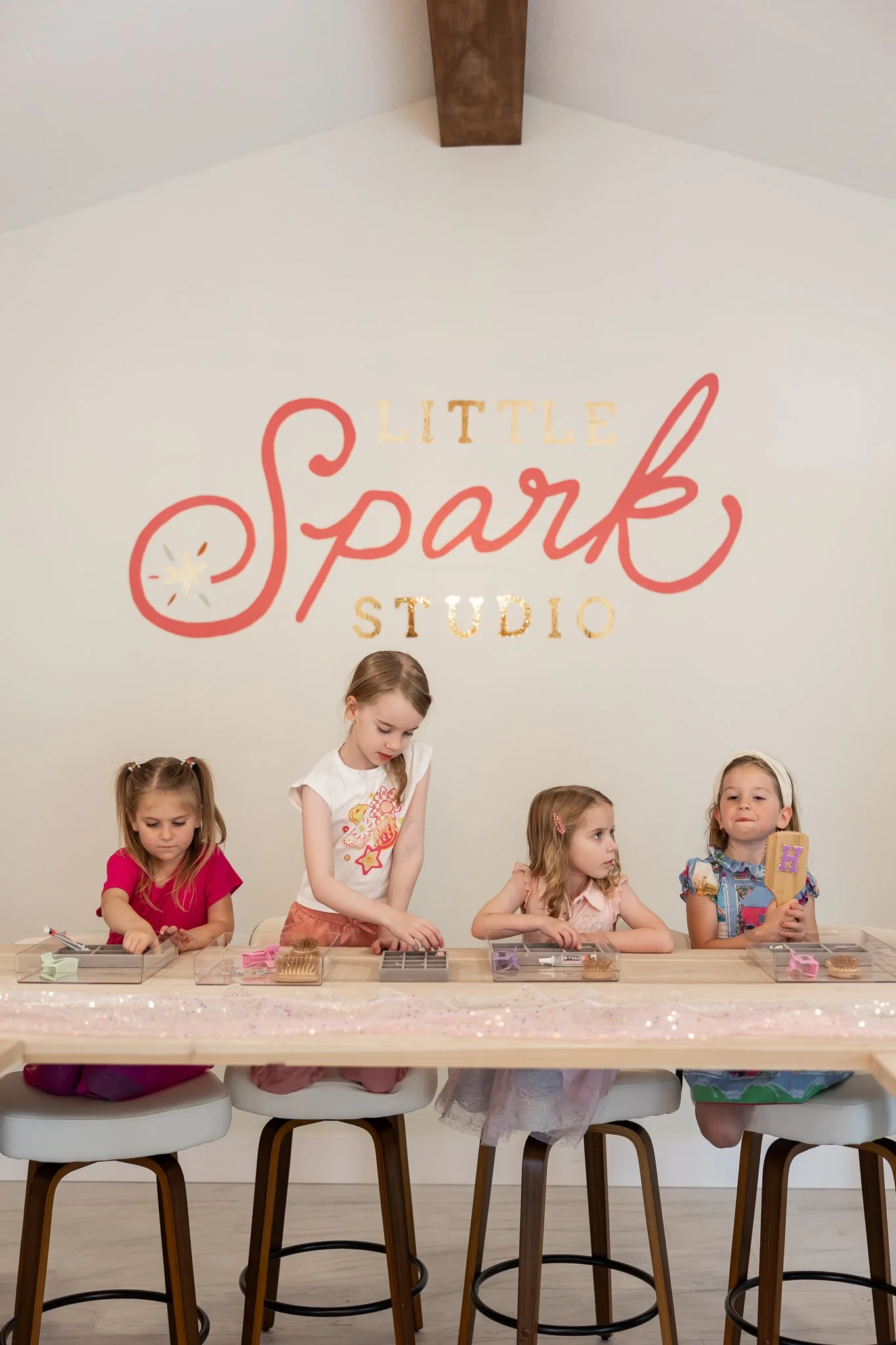 Five young girls participating in a crafting activity at a 'Little Spark Studio' with a sign on the wall behind them, seated at a table with various craft supplies.