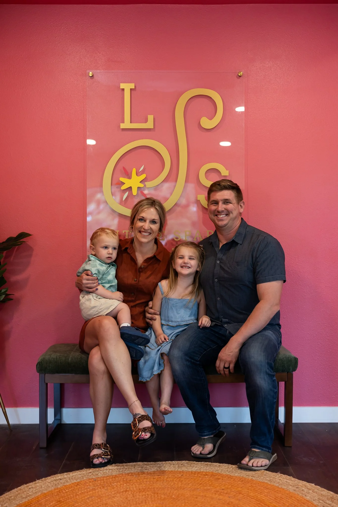 Family of four including a woman, man, young girl, and toddler boy sitting on a bench in front of a pink wall with a signage that reads 'Lush'.