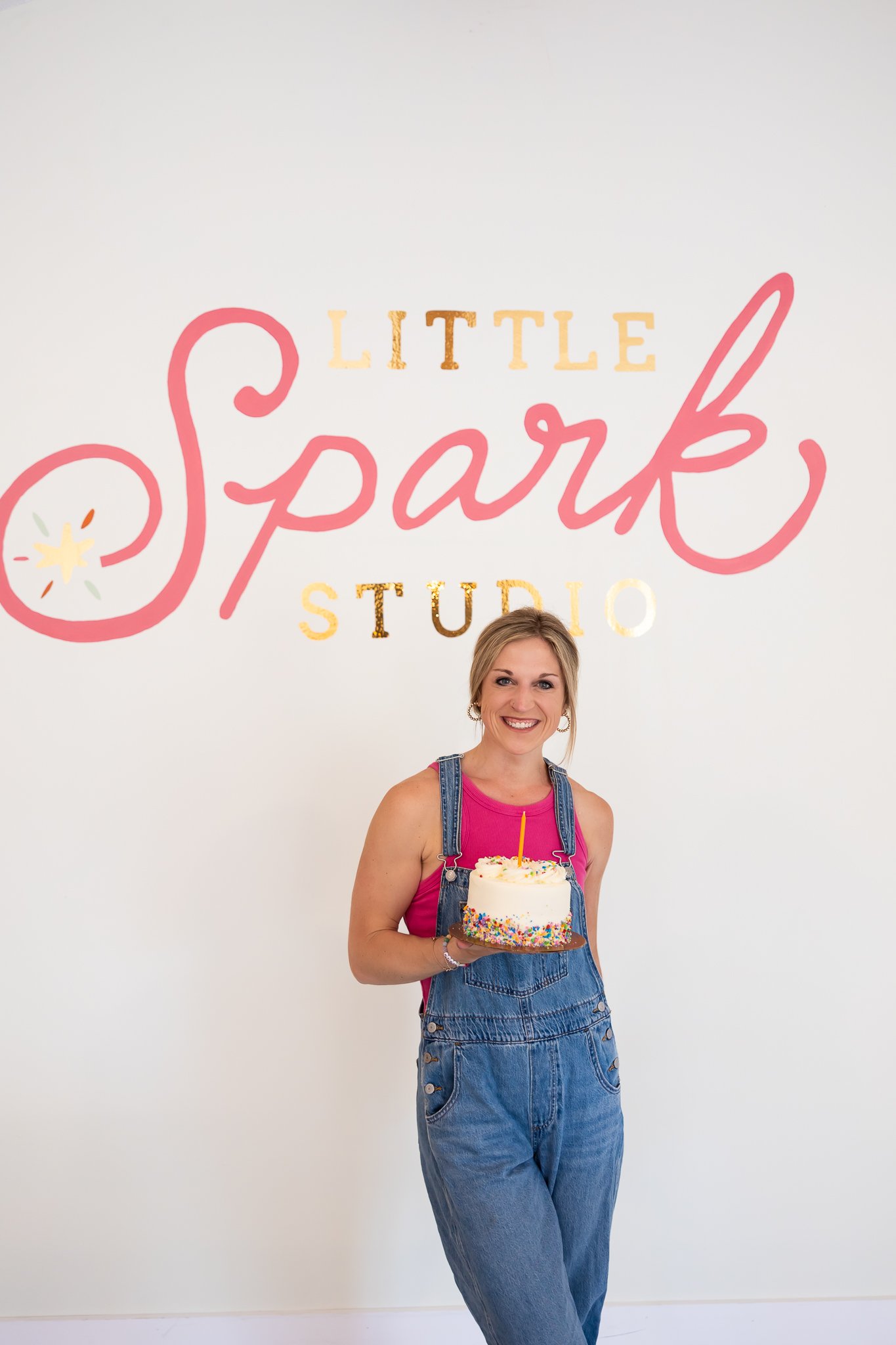 A woman holding a birthday cake with colorful sprinkles, standing in front of a sign that reads 'Little Spark Studio' with a smile.