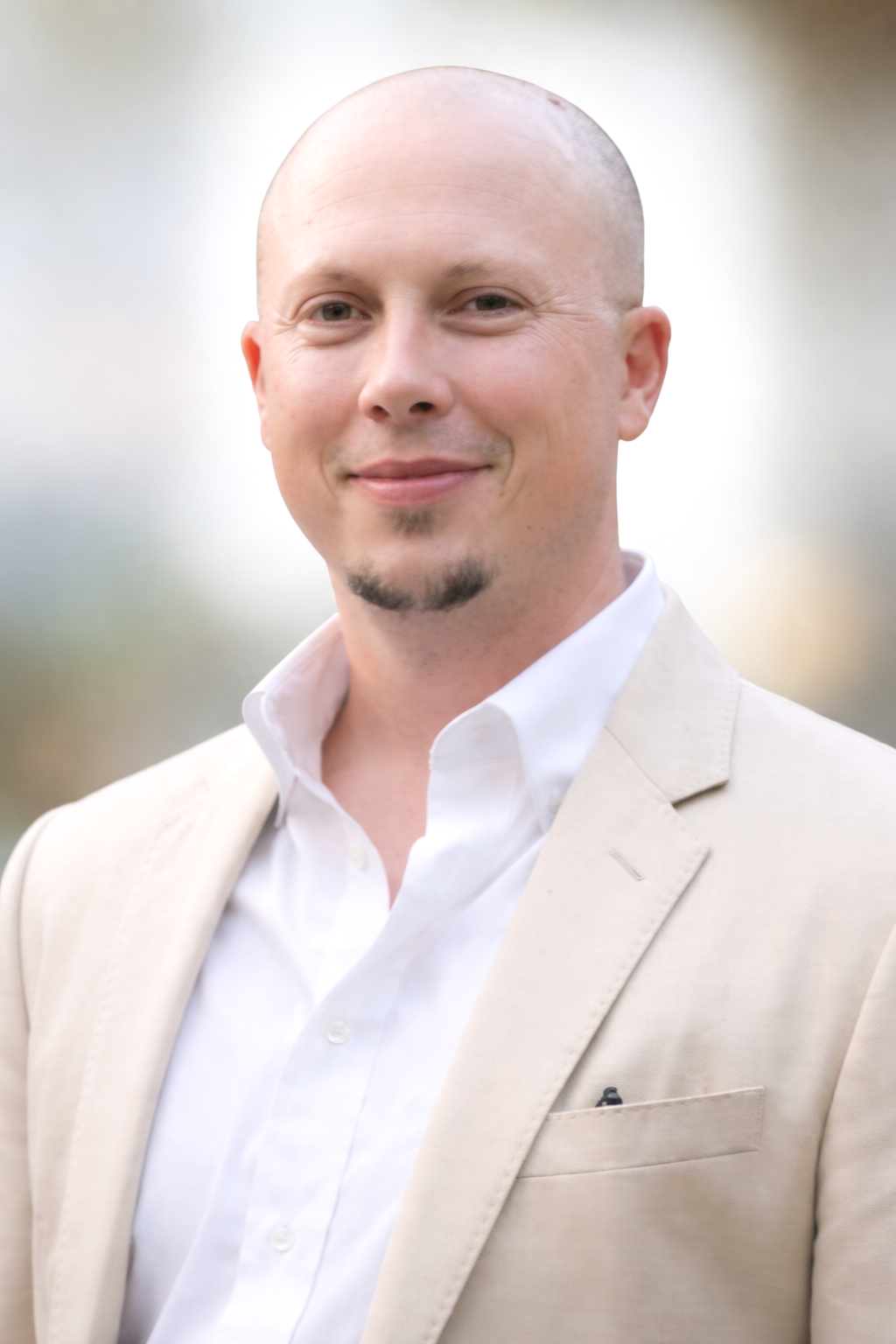A smiling man with a shaved head and small beard in a light beige suit and white shirt, outdoors with blurred natural background.